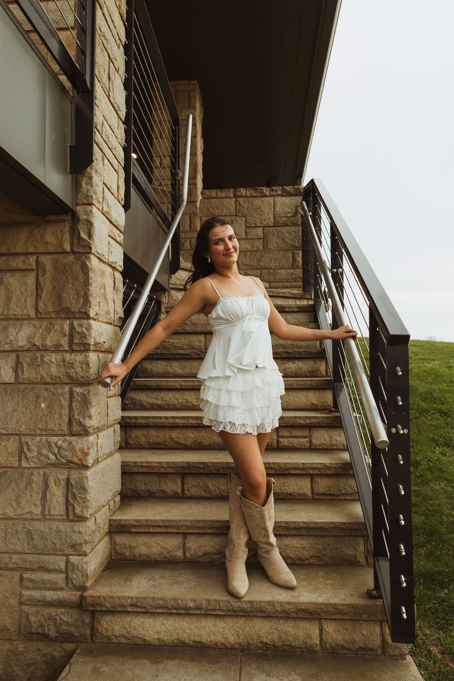 Young woman in white ruffled dress and cowboy boots posing on stone stairs with metal railing.