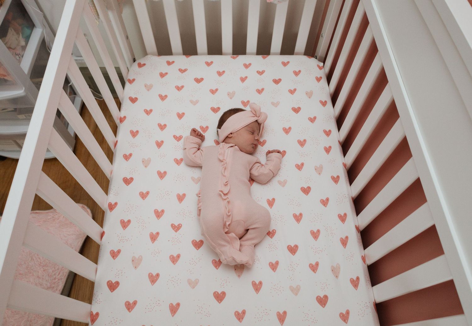 A sleeping baby in a white crib on a pink heart patterned sheet lies peacefully under natural lighting.