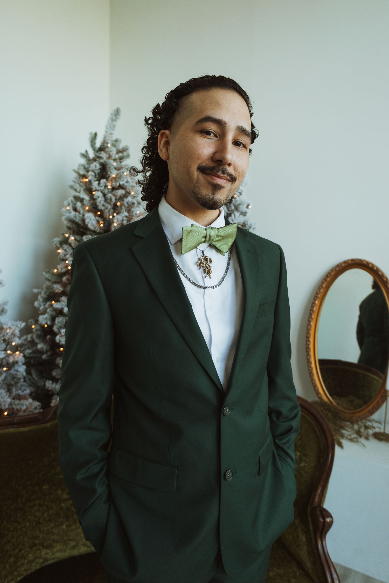Handsome groom in green suit with sage bow tie and brooch posing beside Christmas tree.