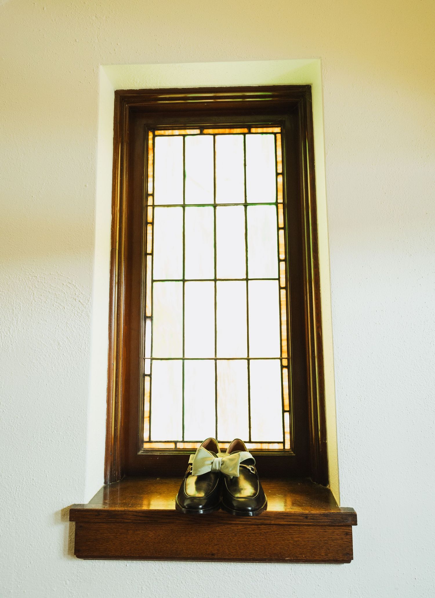 Elegant black dress shoes with bow tie placed on wooden ledge of stained glass church window.