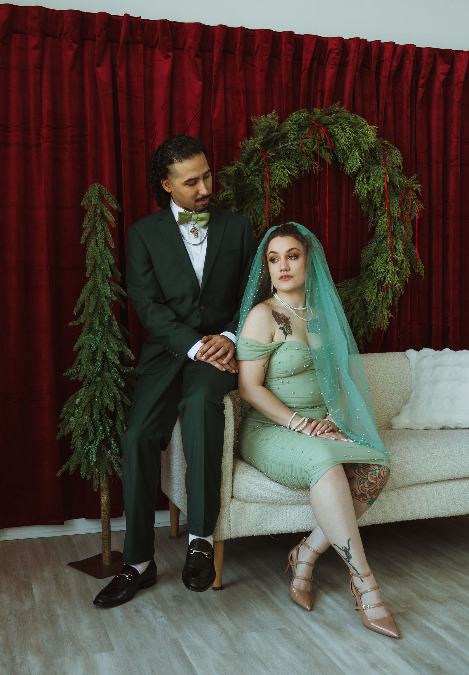 A couple poses in holiday wedding attire, bride in green dress with teal veil beside groom in black suit.