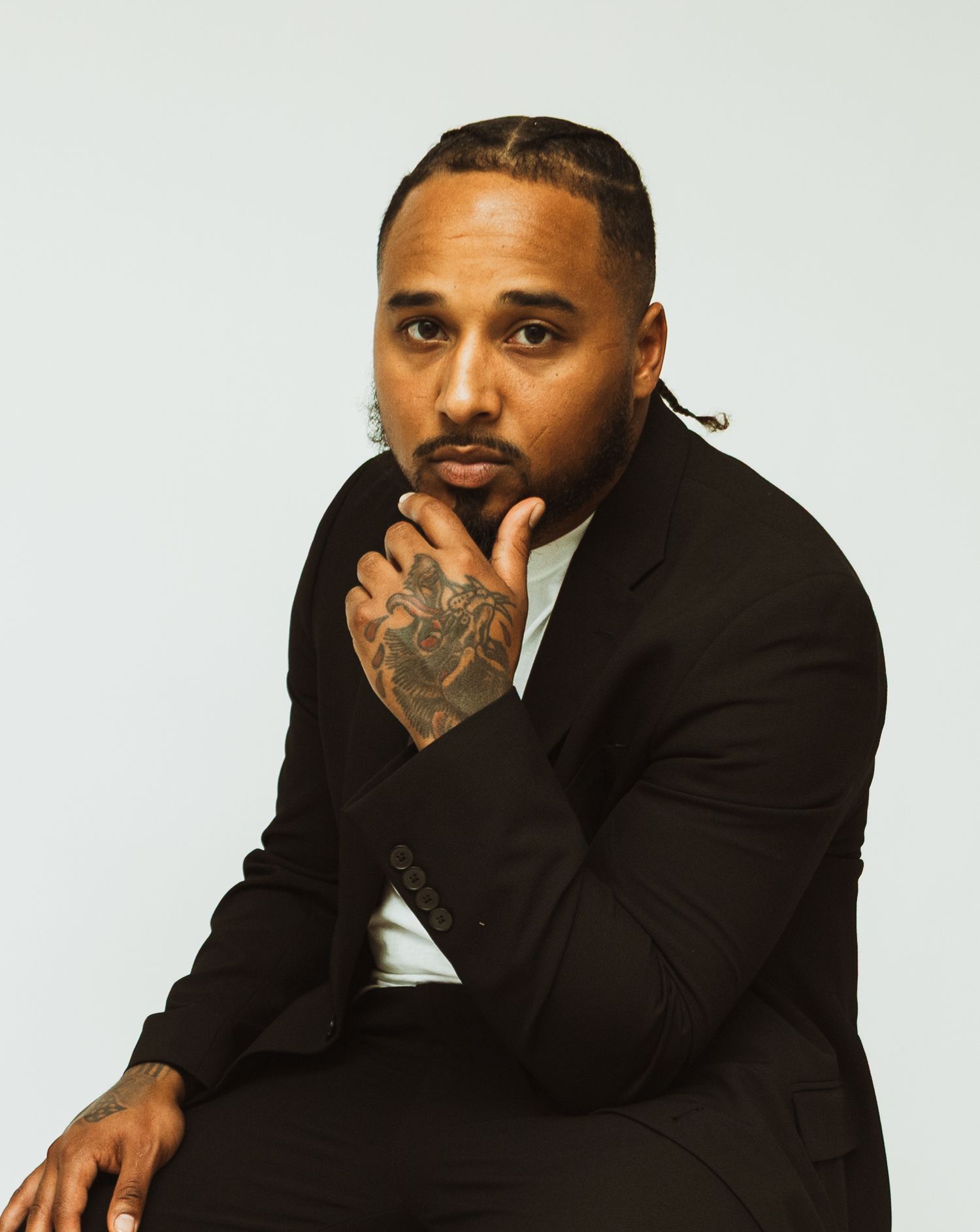 Person wearing a black suit with visible tattoos poses thoughtfully against a light background.