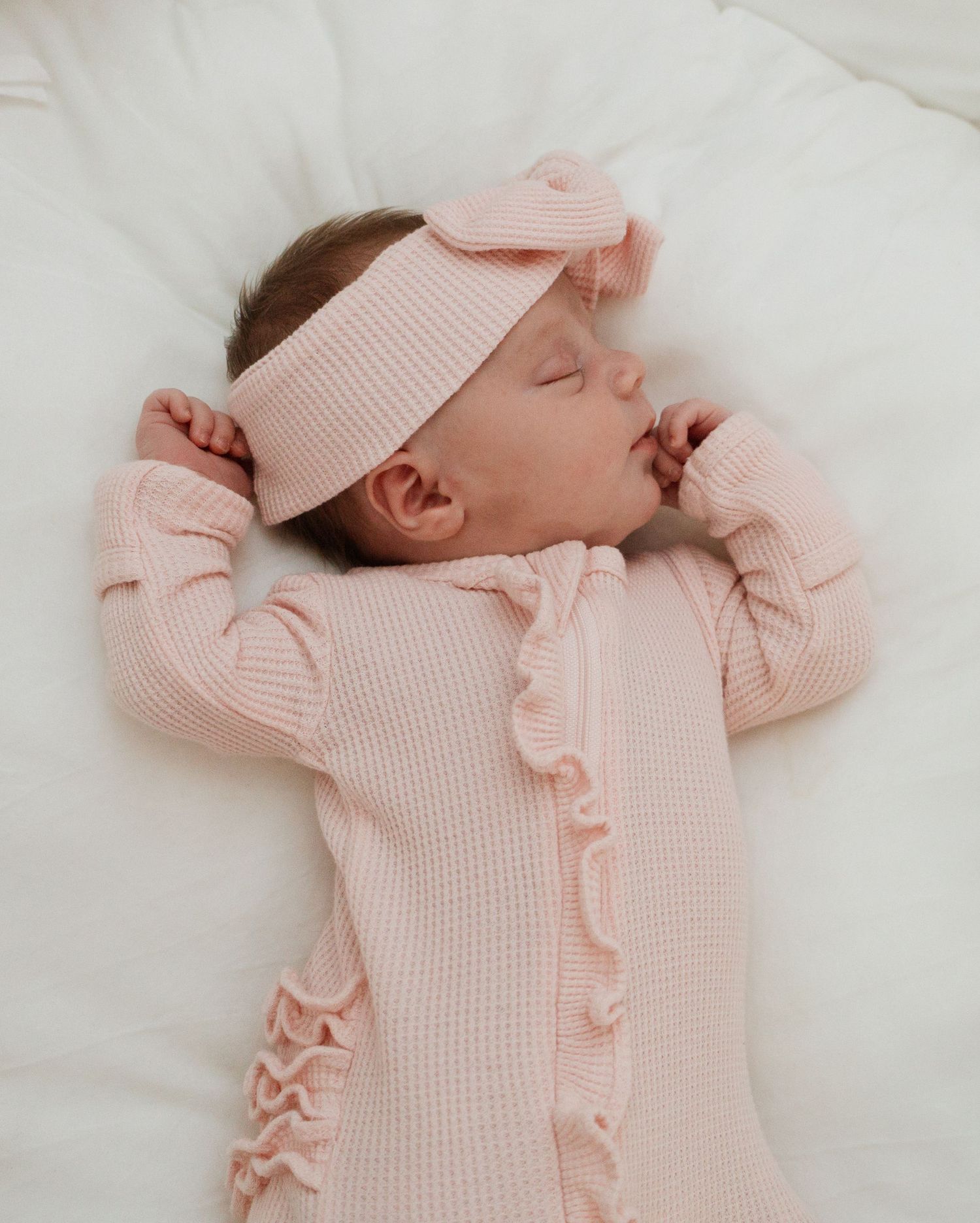 Sleeping baby wearing pink ruffled thermal one-piece and matching headband on white bedding.