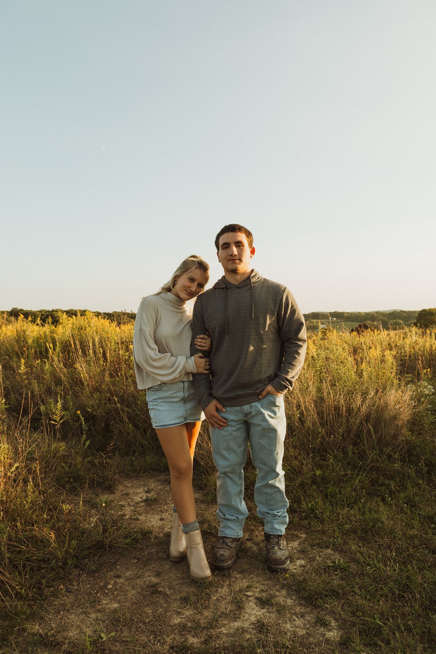 Couple standing together in field during golden hour wearing casual outfits.