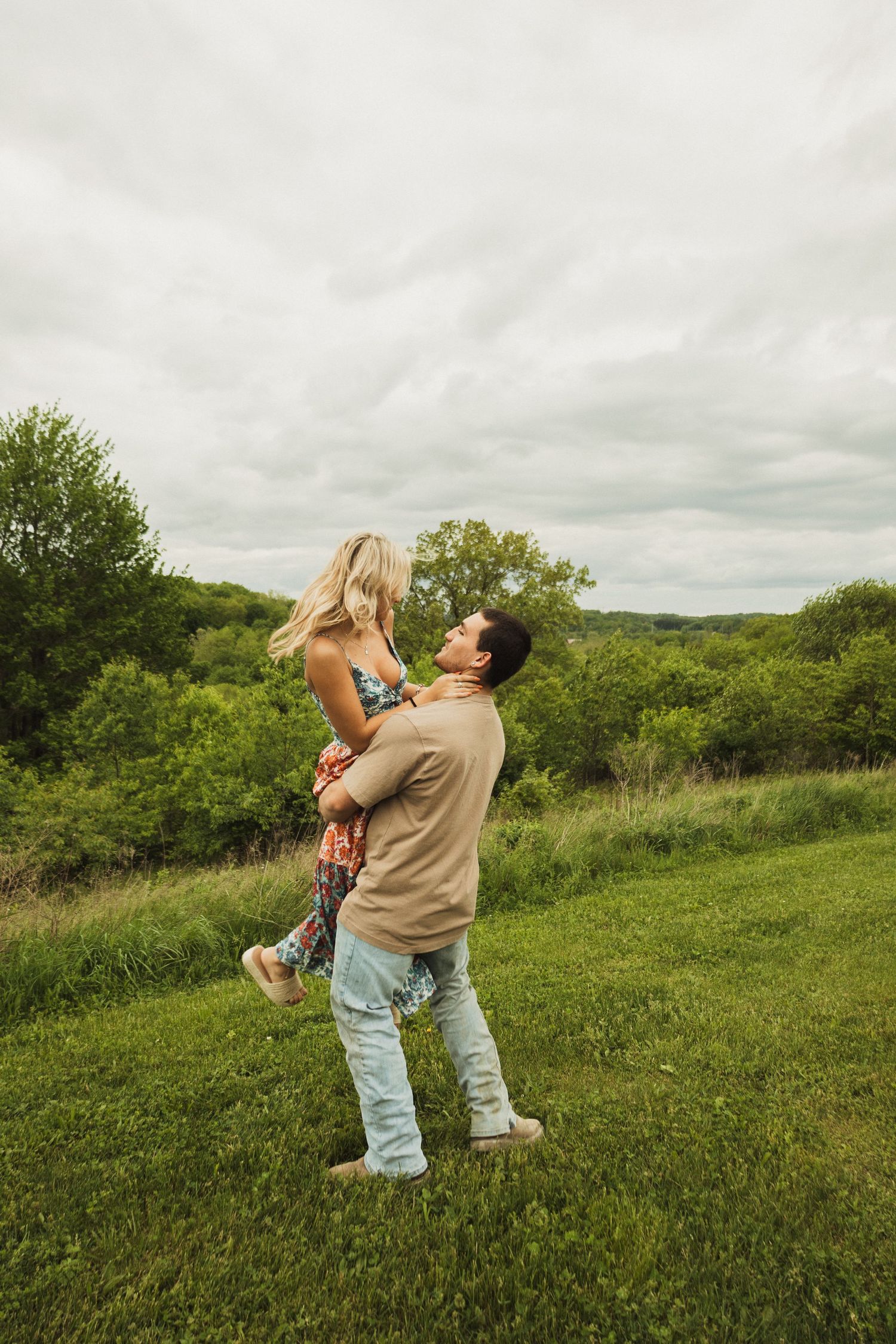 Couple enjoying a playful moment in a grassy field as he lifts her up against a cloudy sky.