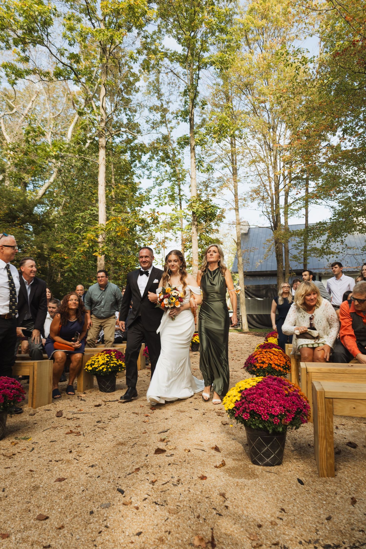 Wedding guests seated on wooden benches during an outdoor autumn ceremony surrounded by trees.