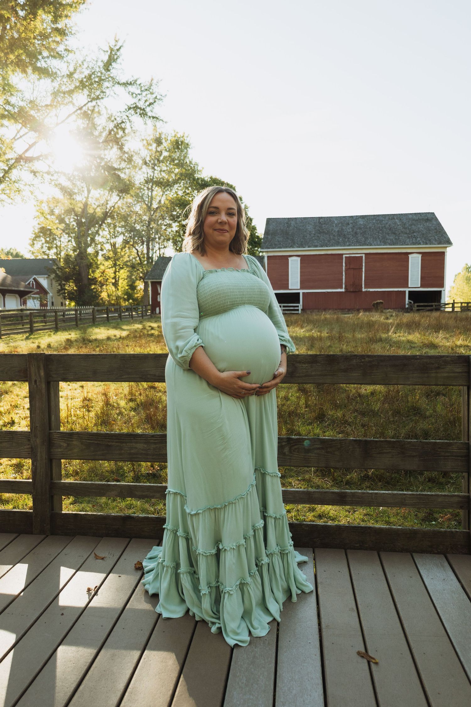A person in a mint green dress poses on a wooden deck with a rustic barn and fence in the background at sunset.