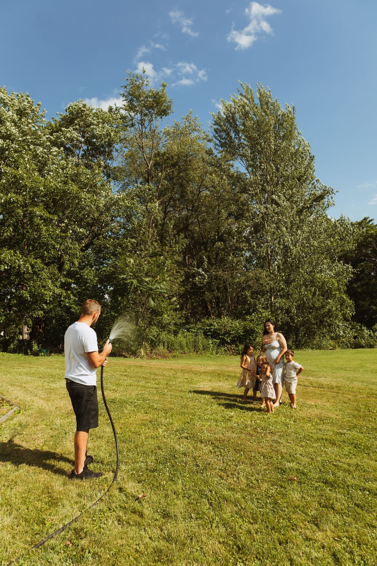 Man spraying water from garden hose at family with children playing on green lawn with trees in background on sunny day.
