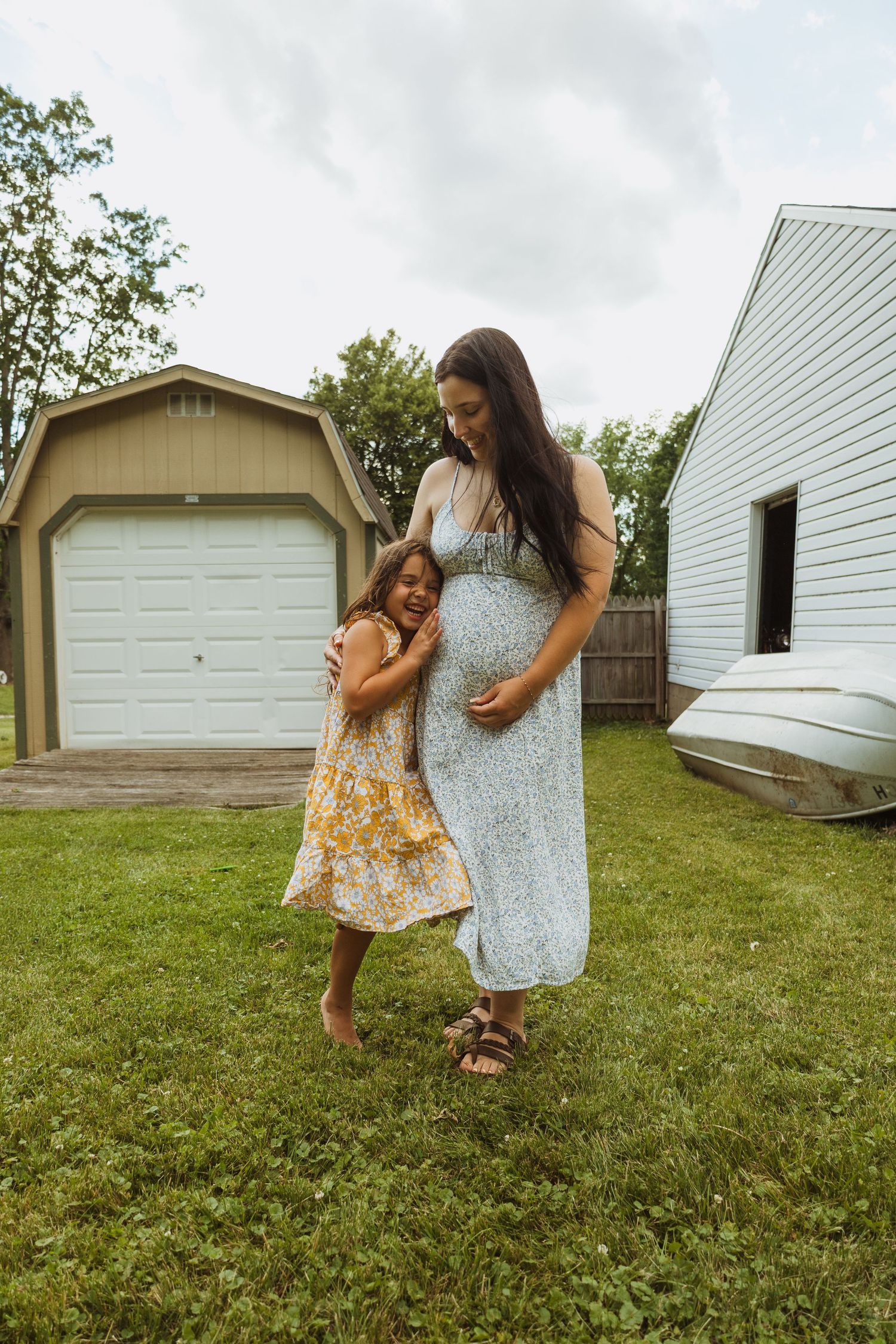Pregnant woman in floral dress with young girl hugging her baby bump in a backyard setting.