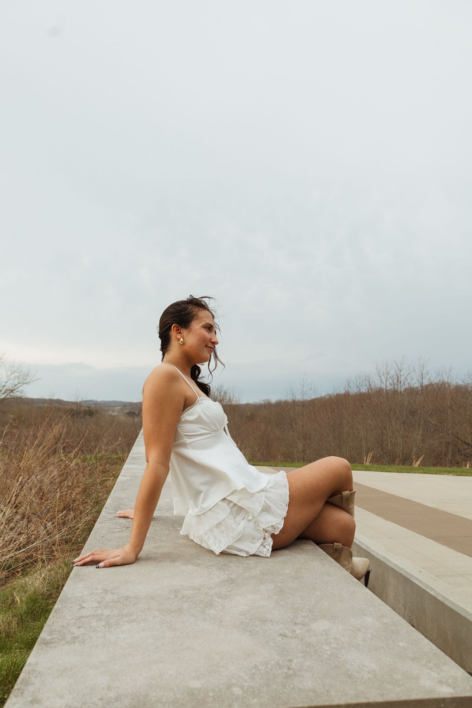 A young woman in a white ruffled mini dress sits on a concrete ledge overlooking a scenic countryside landscape.