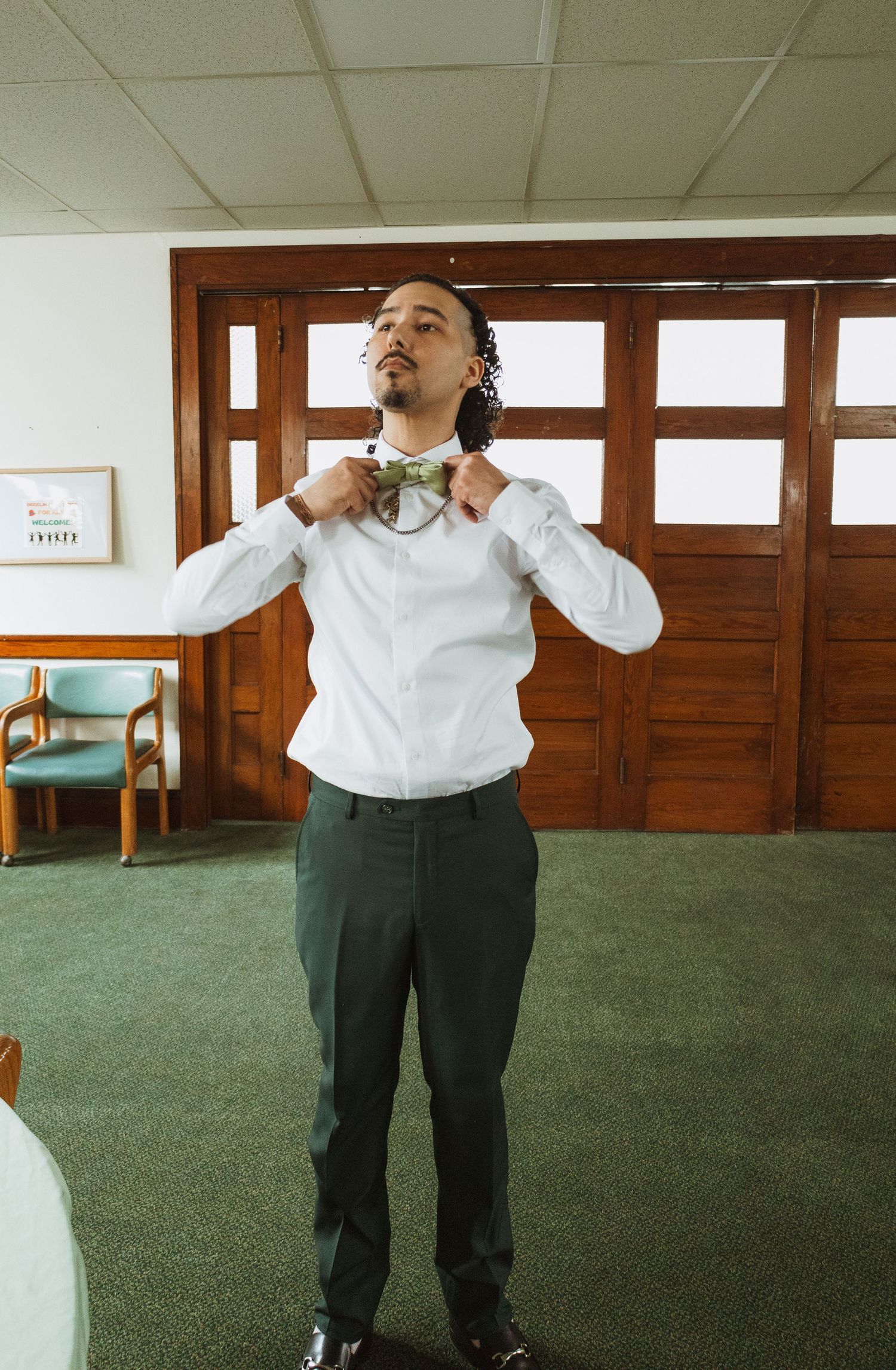 Groom adjusting bow tie while getting ready in white shirt and dark trousers in a carpeted room.