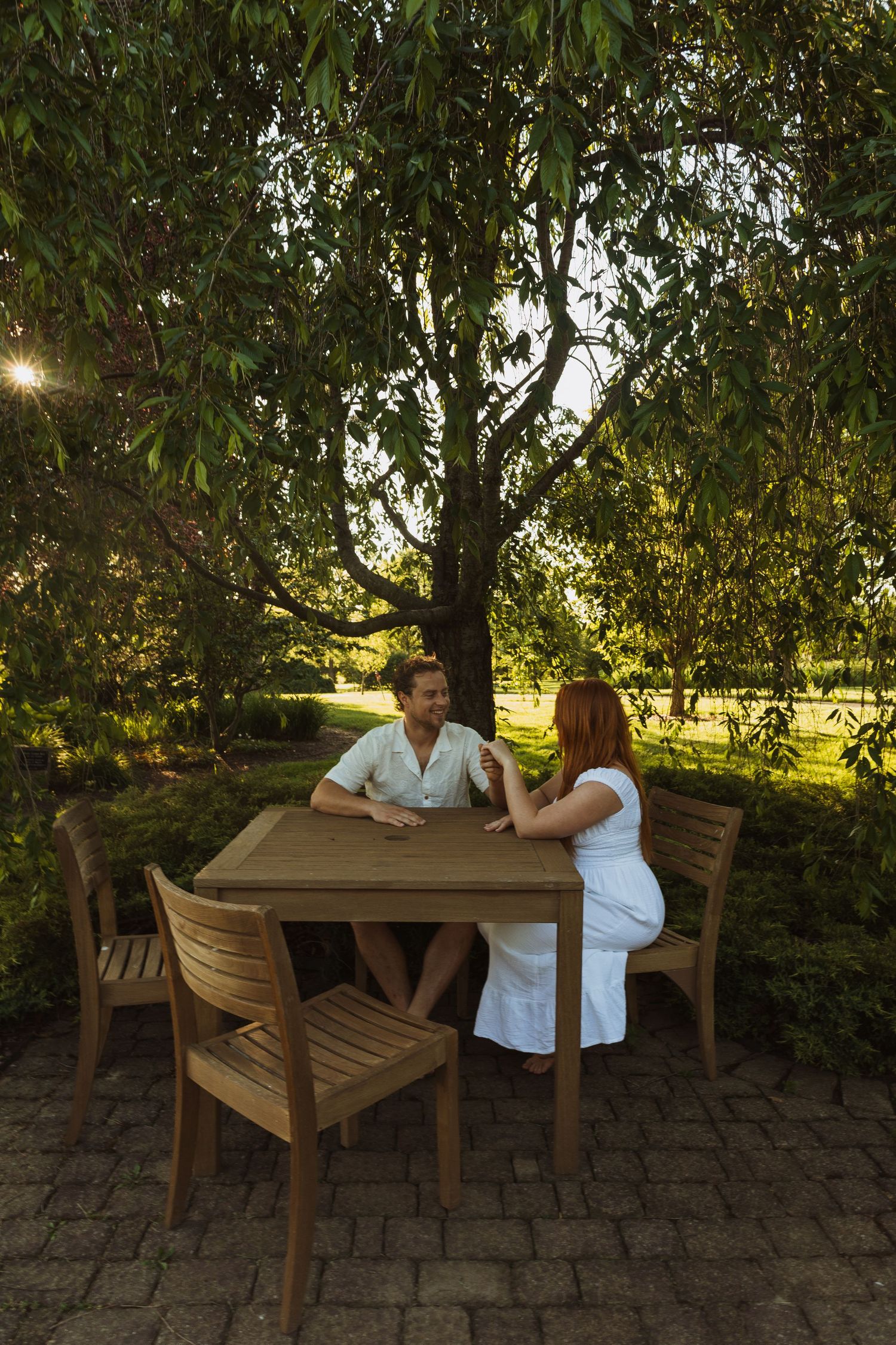Couple sitting at a wooden outdoor table under a lush green tree during golden hour sunset in a garden.