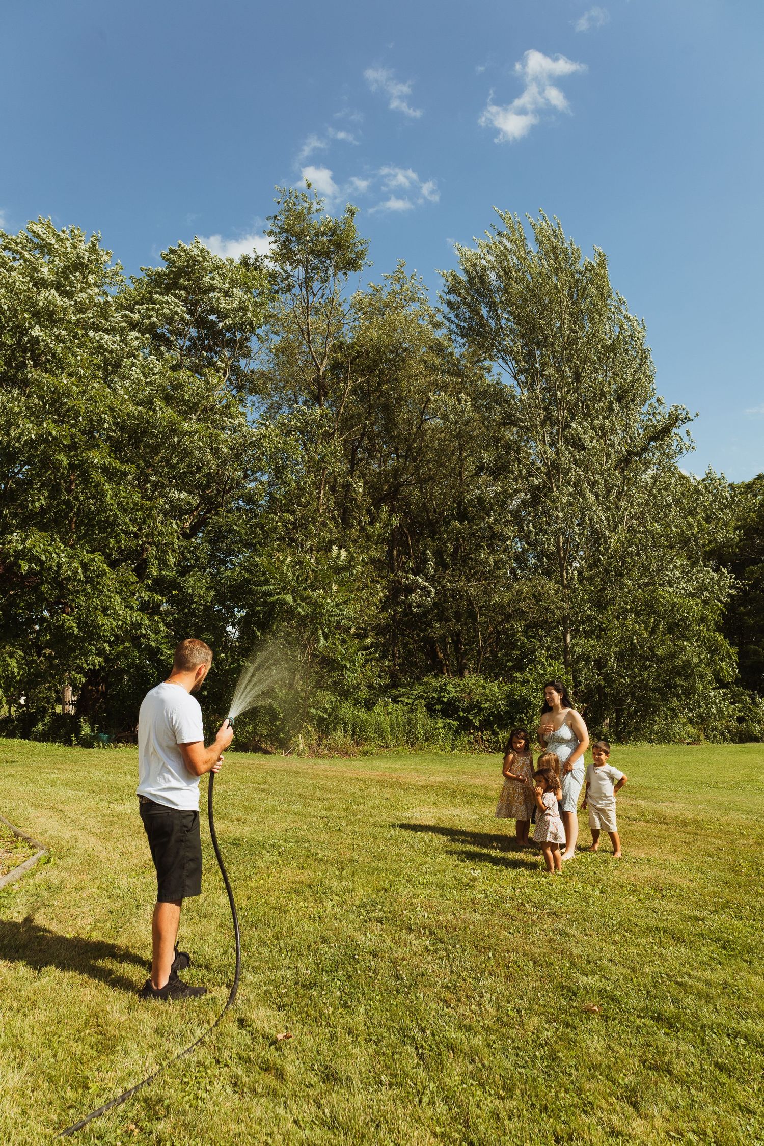 Family enjoying summer fun in backyard, man spraying water hose at woman and children on green lawn.