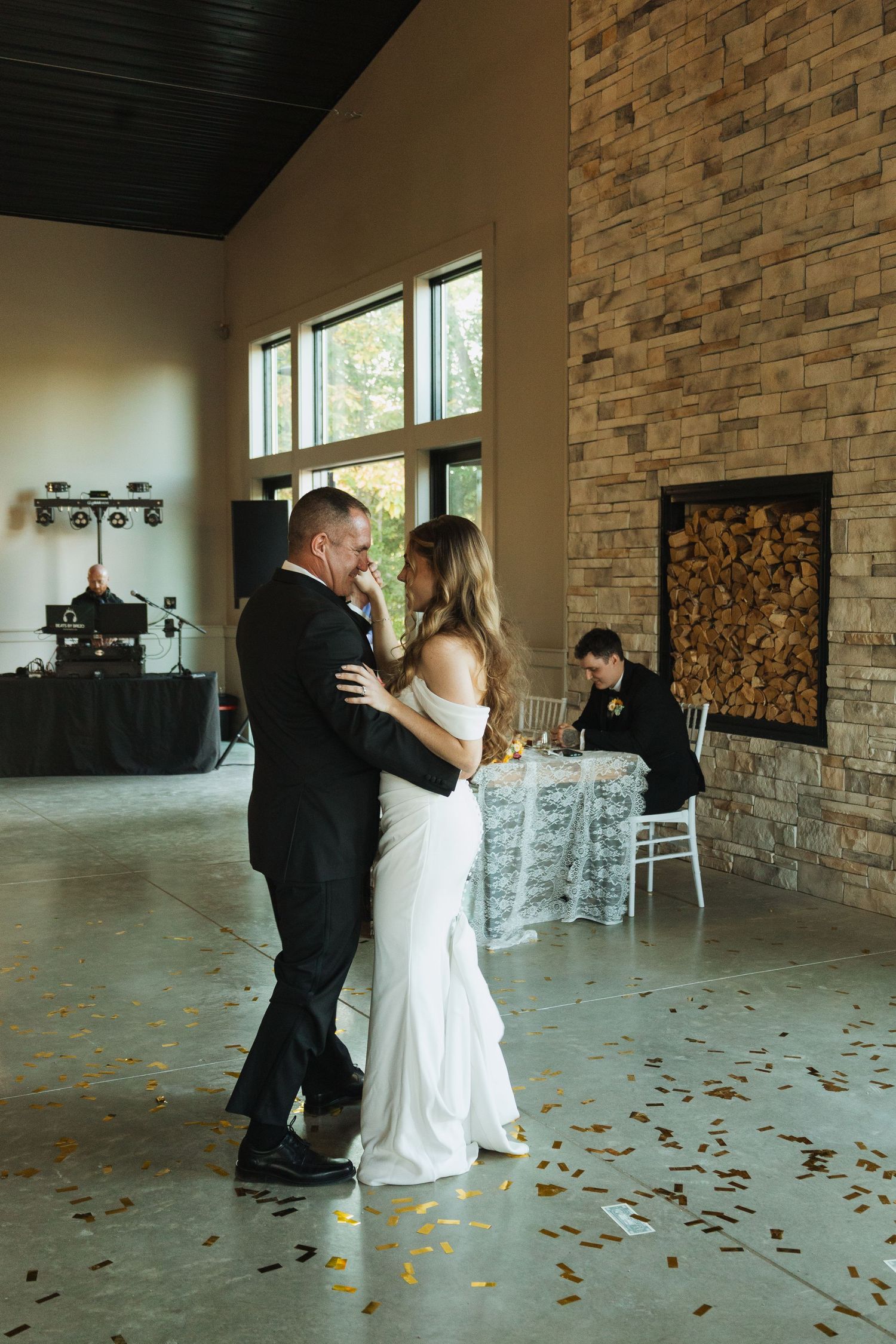A couple shares their first dance in a rustic venue with brick walls and scattered rose petals on the floor.
