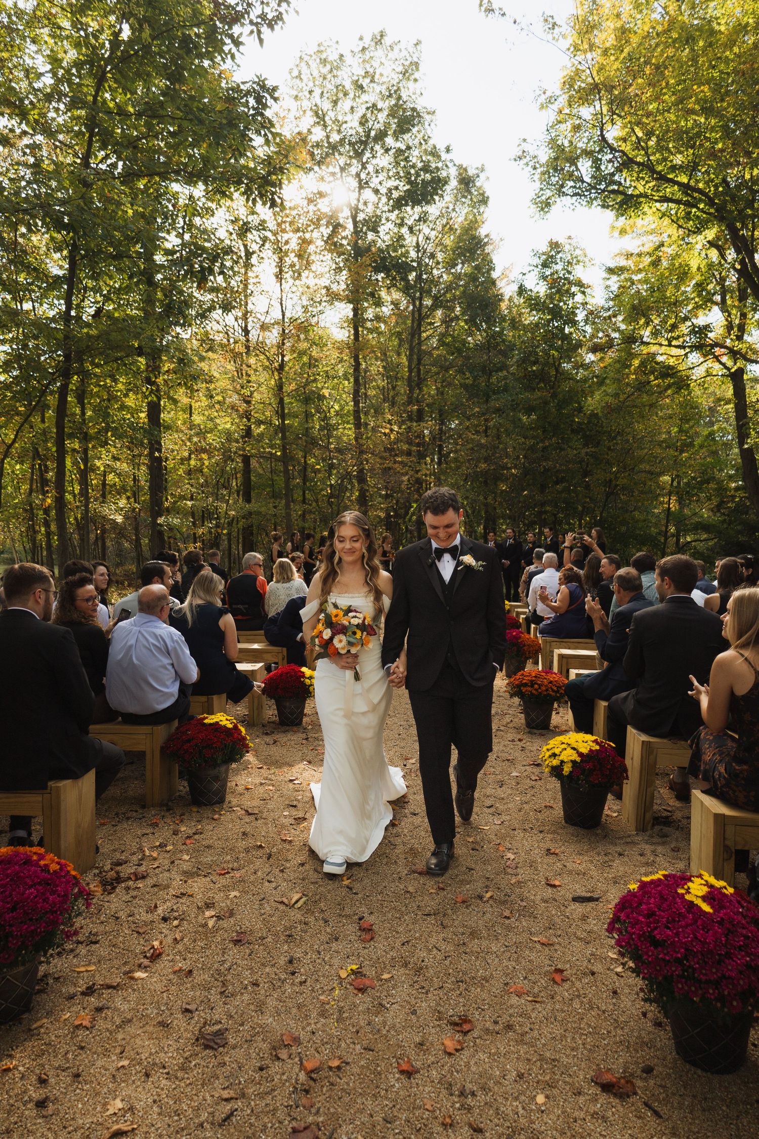 Outdoor forest wedding ceremony with wooden bench seating and burgundy mum decorations.