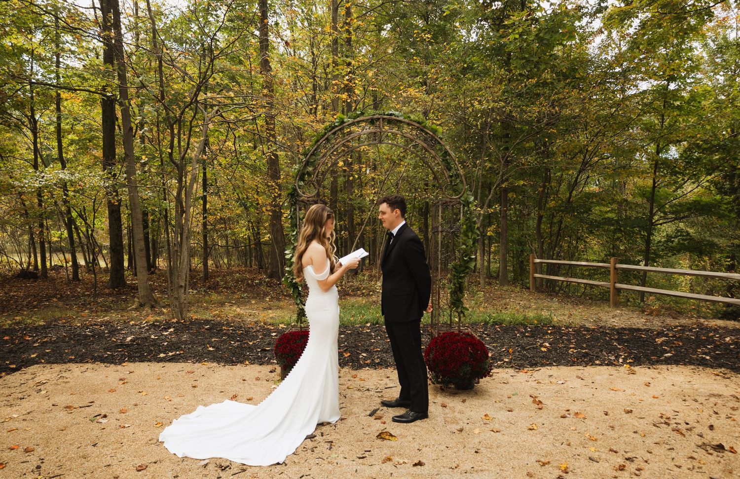 Wedding ceremony under autumn trees with red mums decorating sandy path.