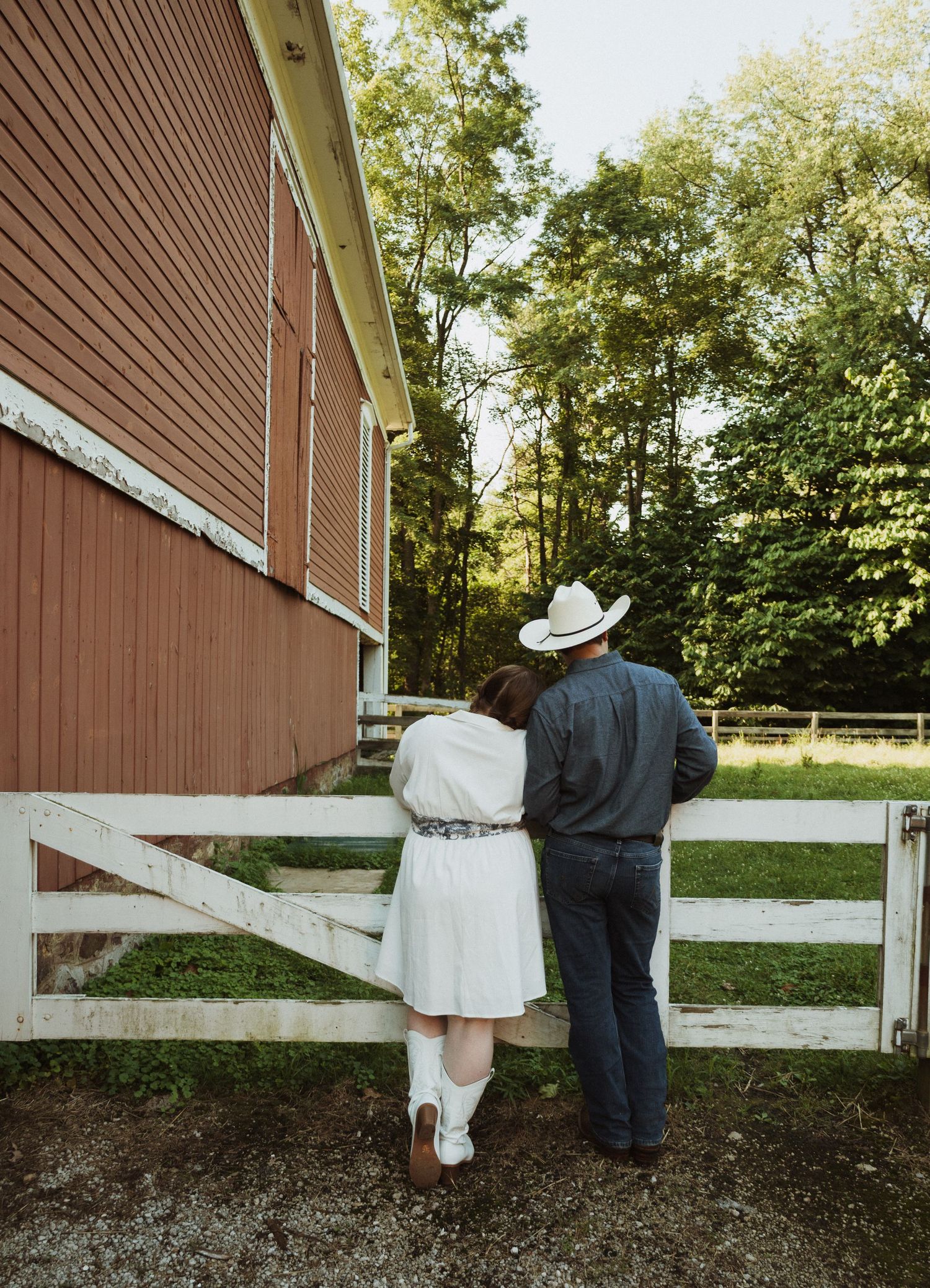 Two people lean against a white wooden fence in front of a brown barn on a sunny day.