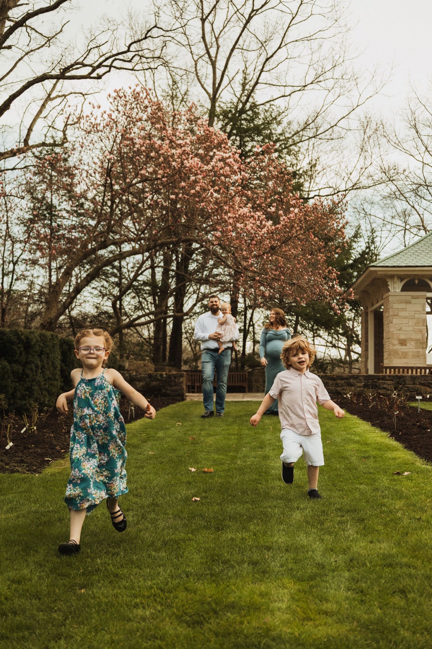 Children running and playing on a grassy path with blooming cherry trees and a gazebo in the background.