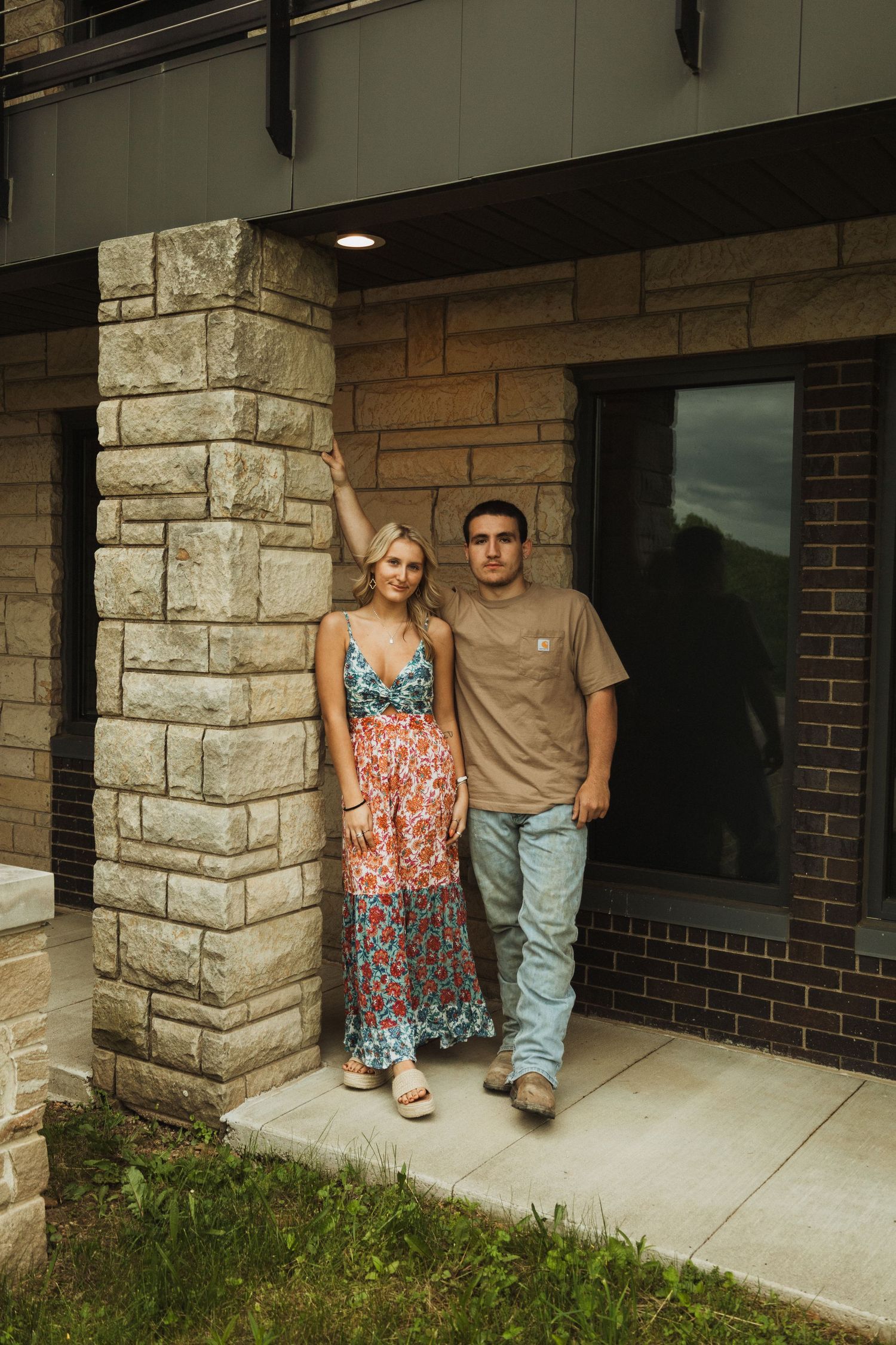 A couple poses against a stone pillar outside a modern building entrance.