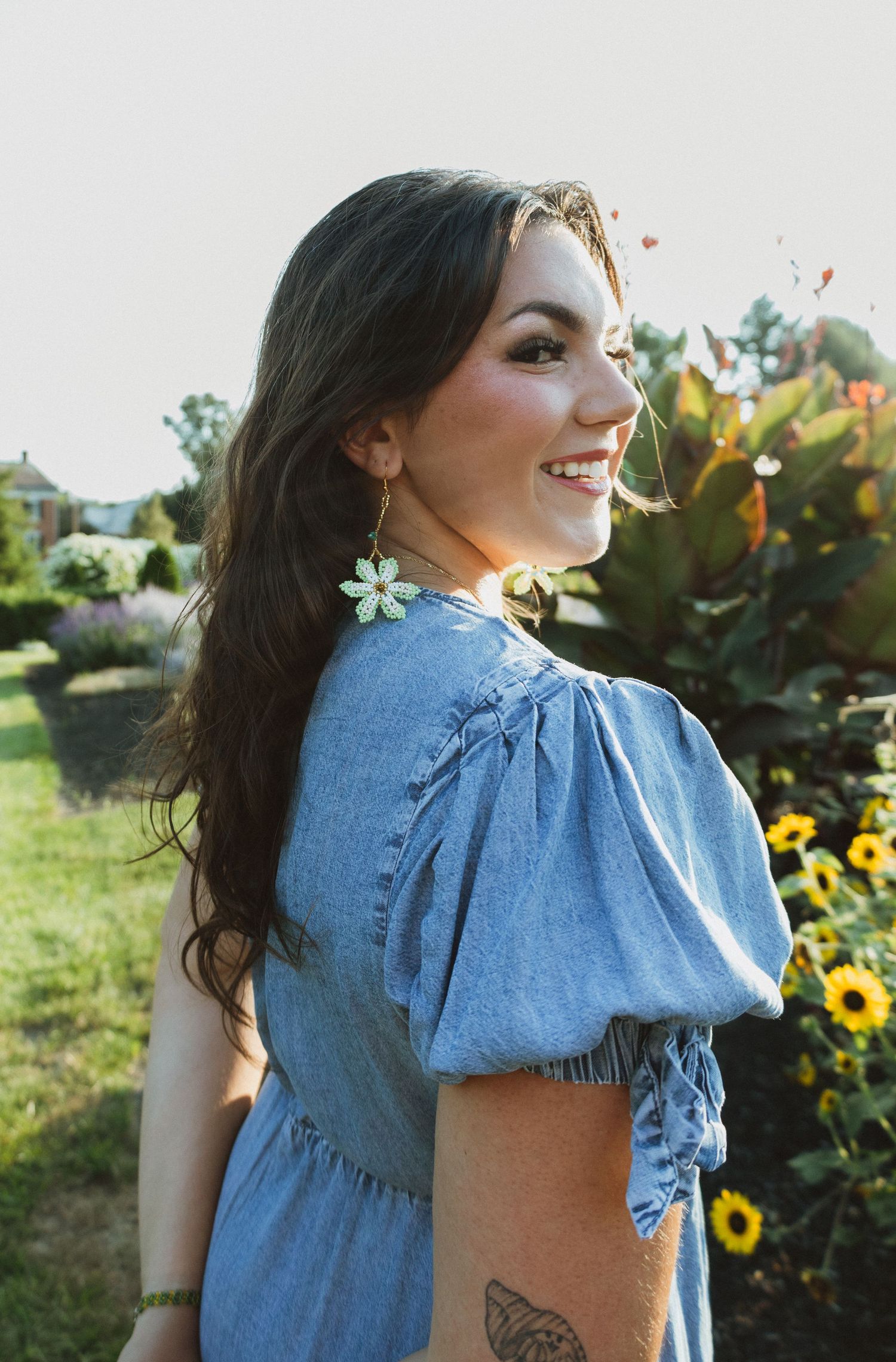 Woman with dark hair wearing a light blue ruffle sleeve dress poses in front of sunflowers in a garden at sunset.