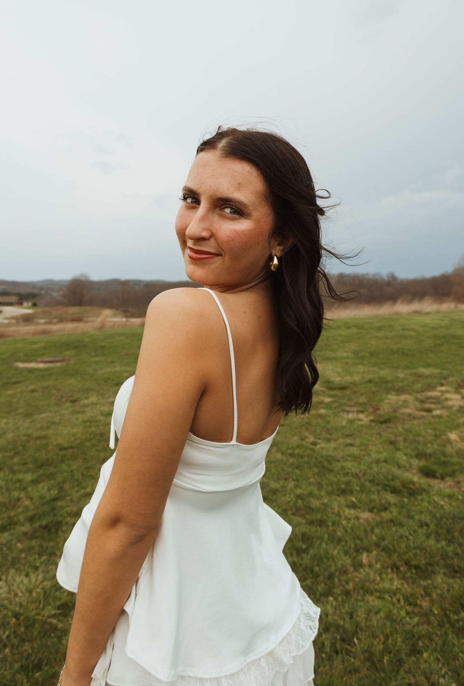 Woman in white dress standing in green field, looking over shoulder with smile, overcast sky background.