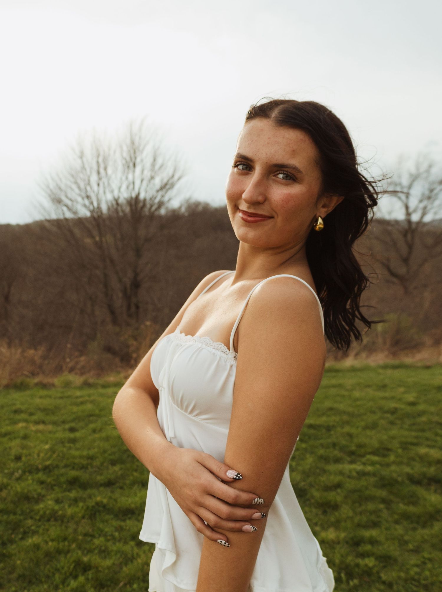 Young woman in white dress posing outdoors on green grass with bare trees in background.