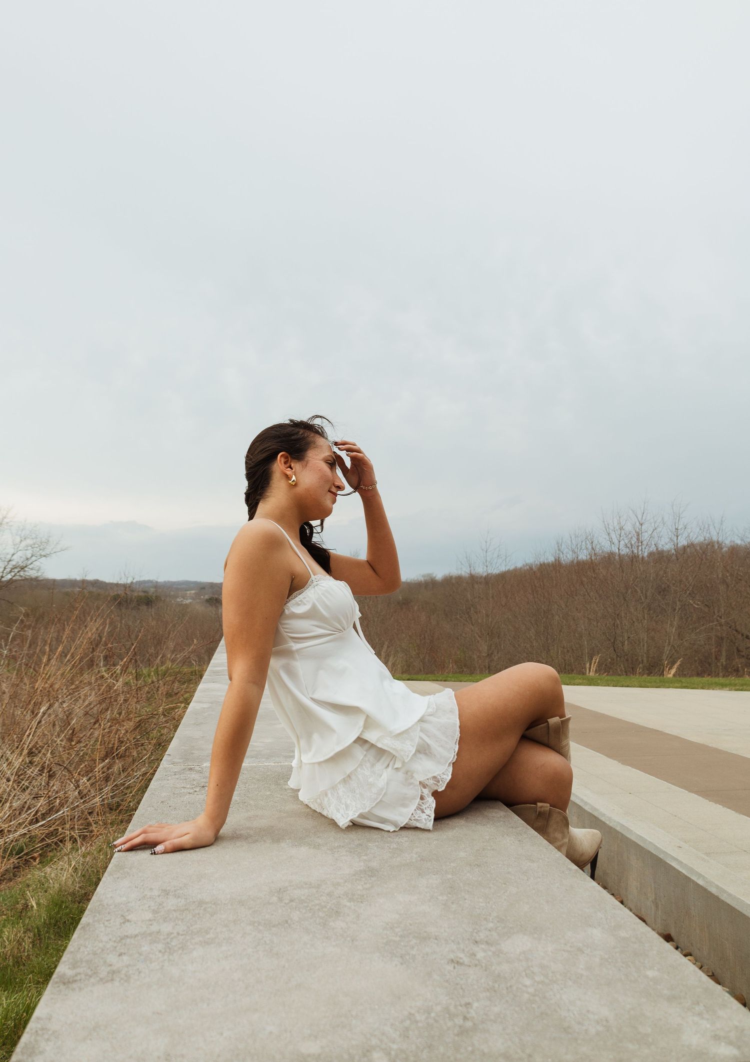Woman in white dress sitting on concrete ledge gazing into distance with overcast sky and autumn landscape behind her.