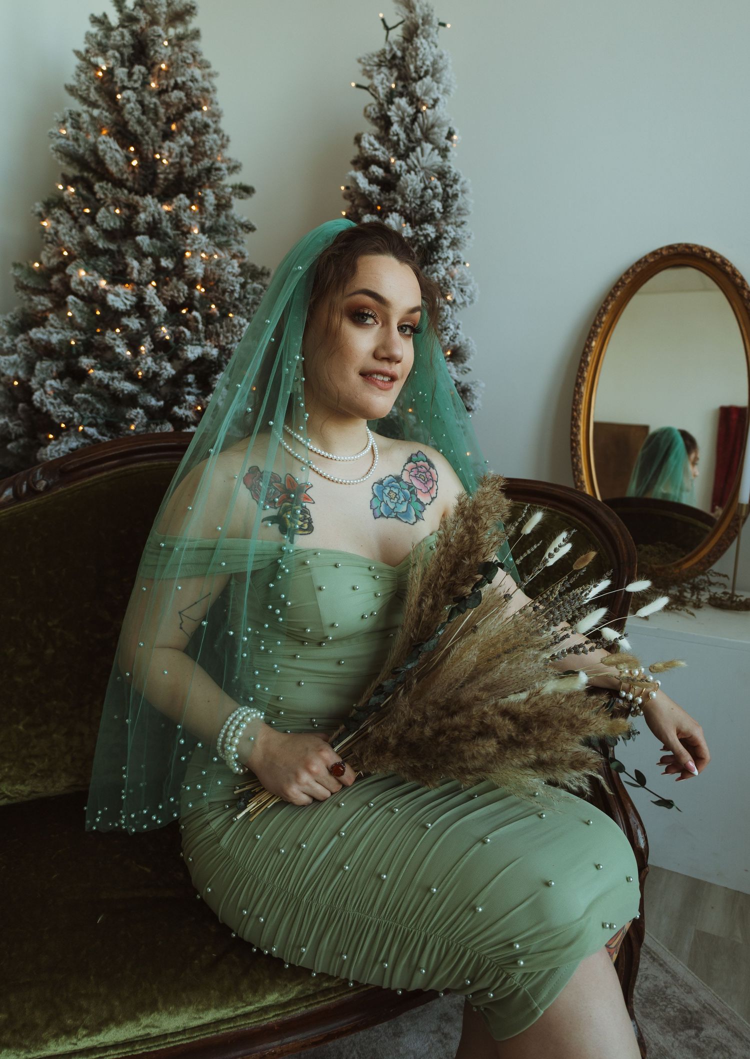 Bride in sage green pearl wedding dress and teal veil holds dried pampas grass by Christmas trees.