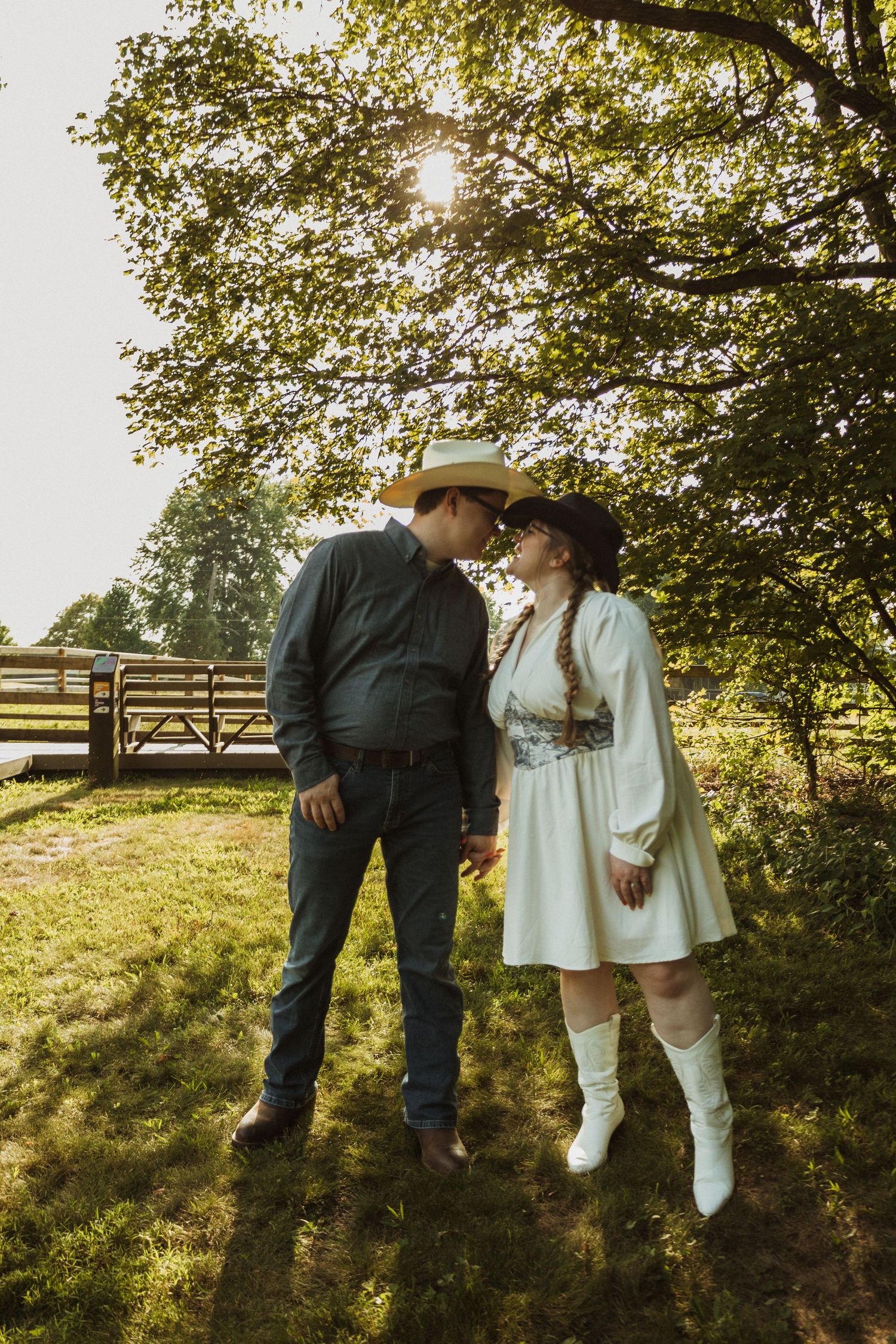 A couple sits together on a wooden fence in a scenic country setting with green trees and grass.