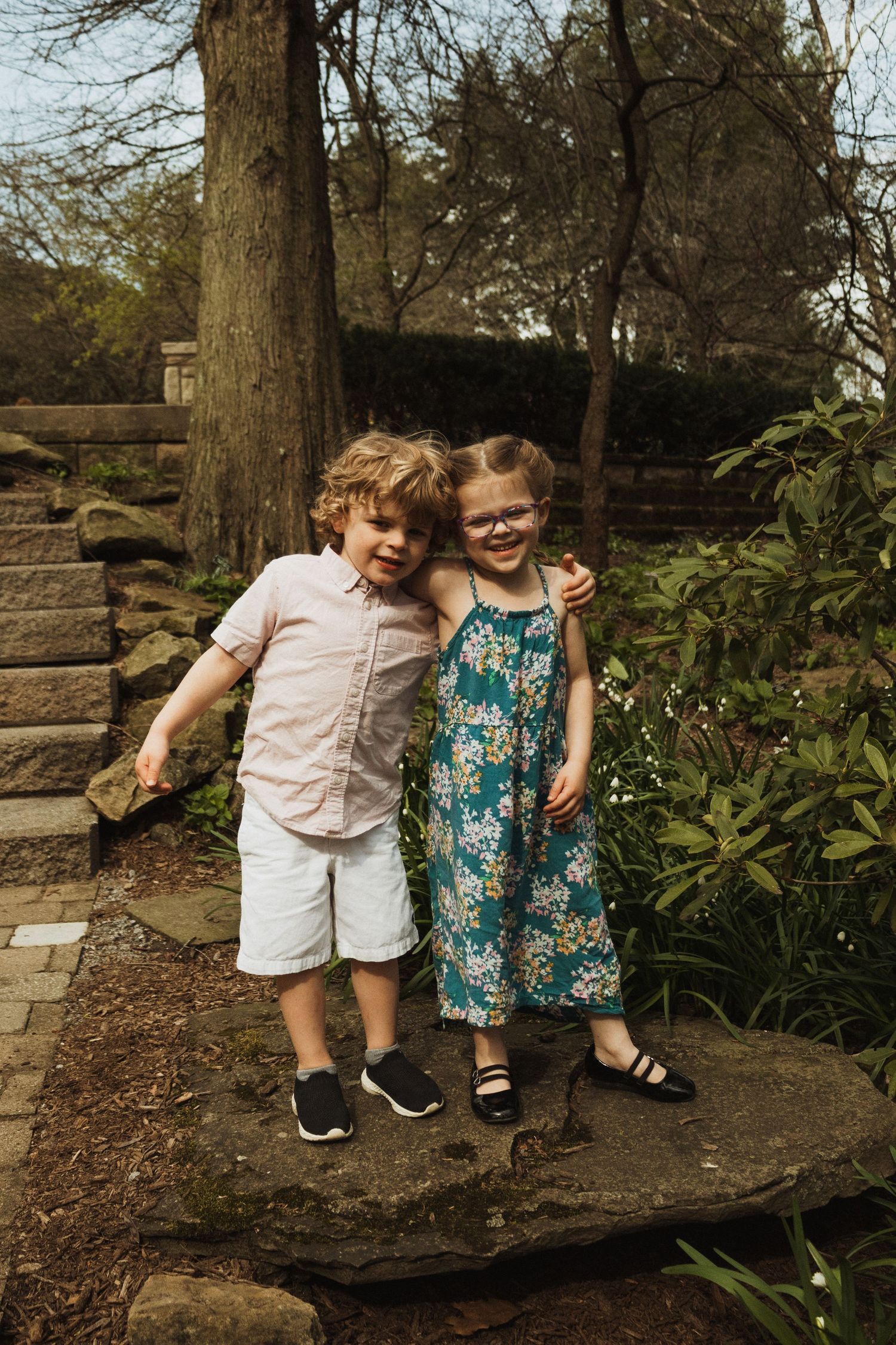 Two young children stand together on a stone path in a garden wearing summer outfits.