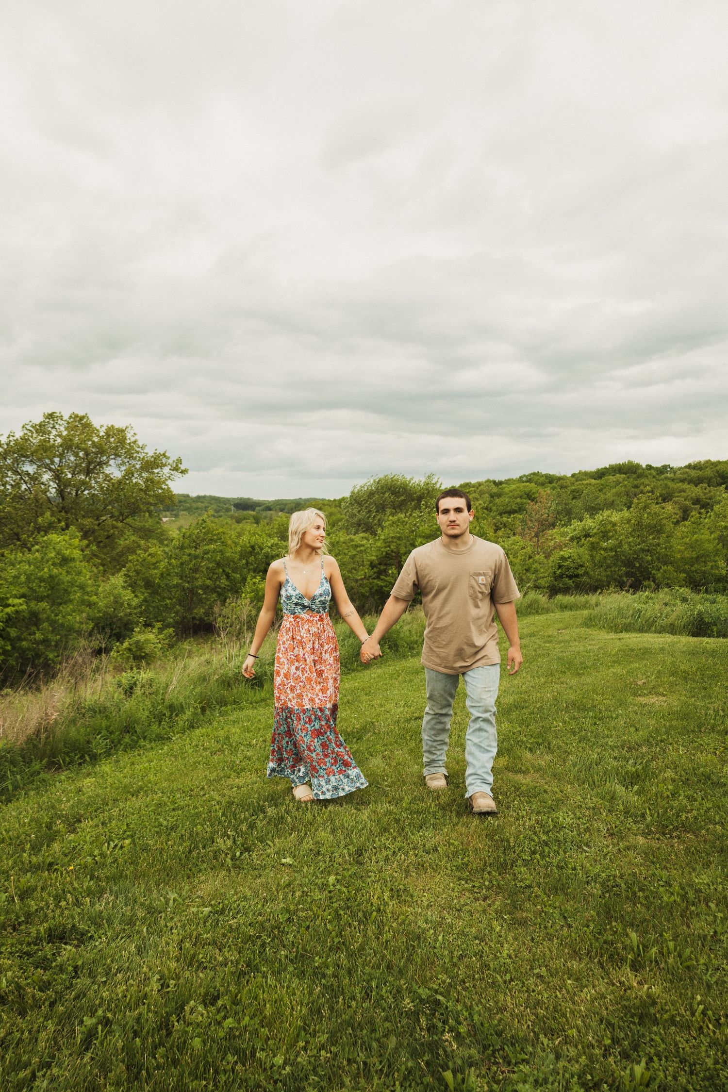 A couple holding hands while walking on a grassy hillside under cloudy skies in a natural landscape setting.