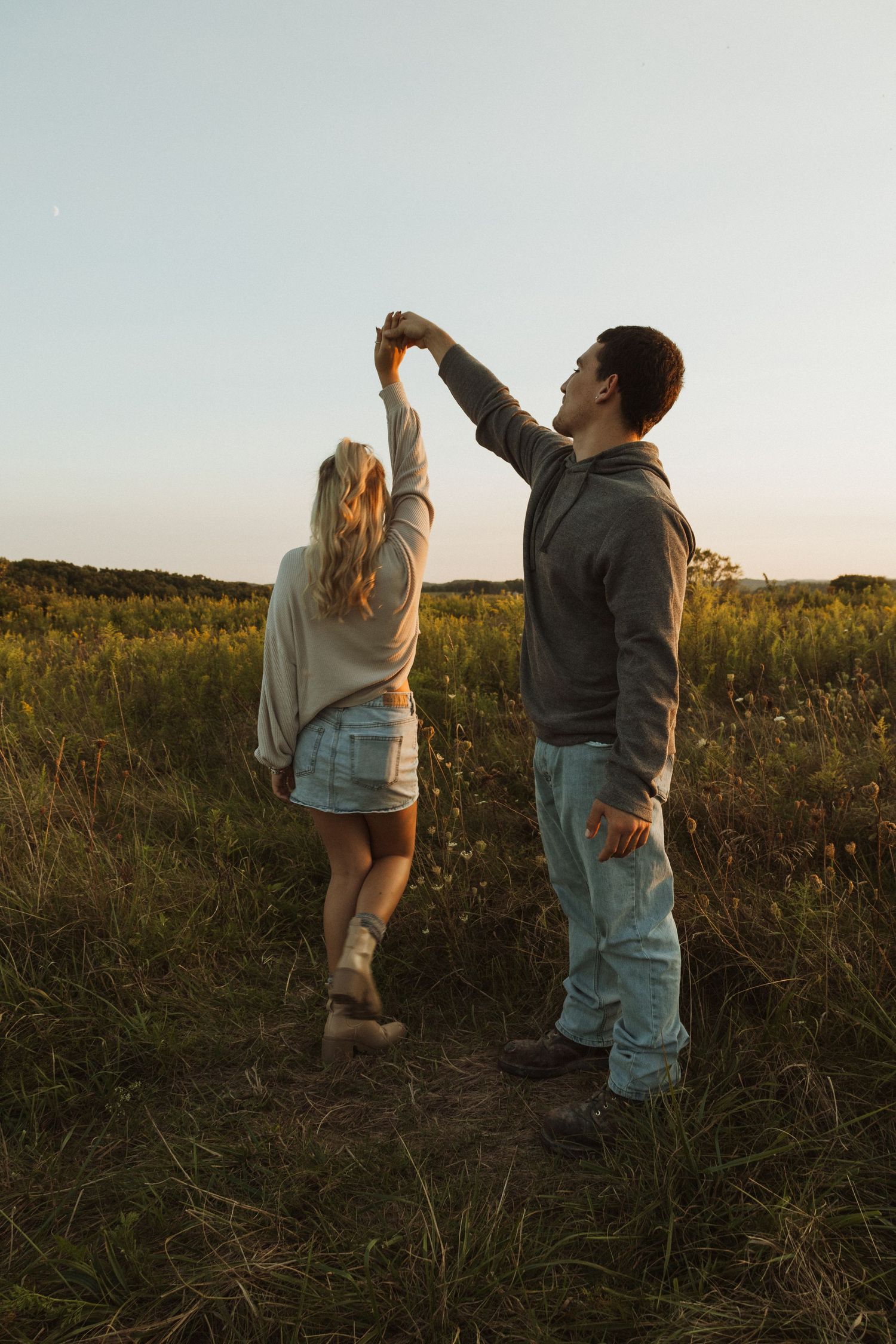 Two people hold hands up towards the sky while standing in a meadow at sunset.
