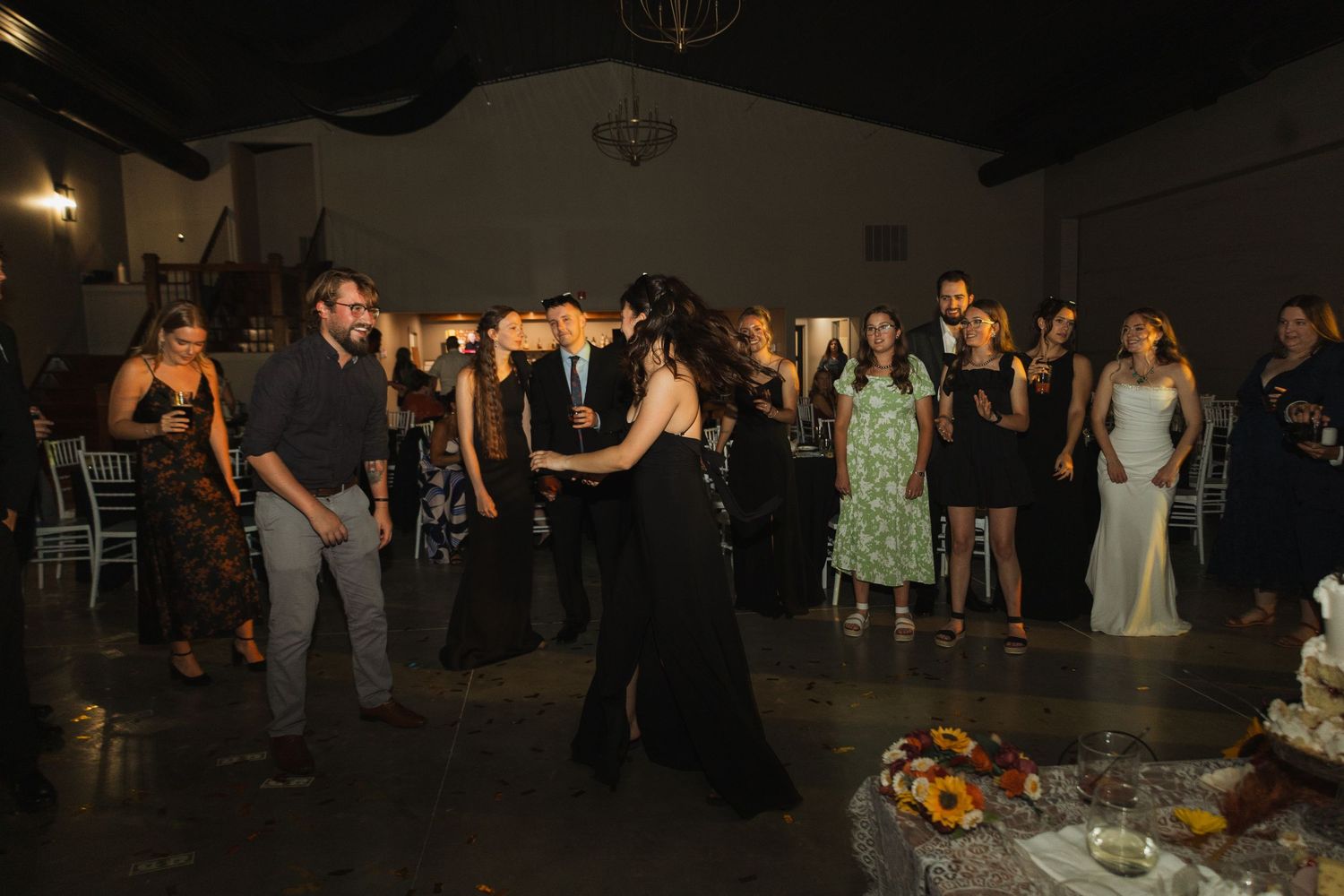 Wedding reception guests dance and celebrate in a dimly lit venue with white chairs and tables.