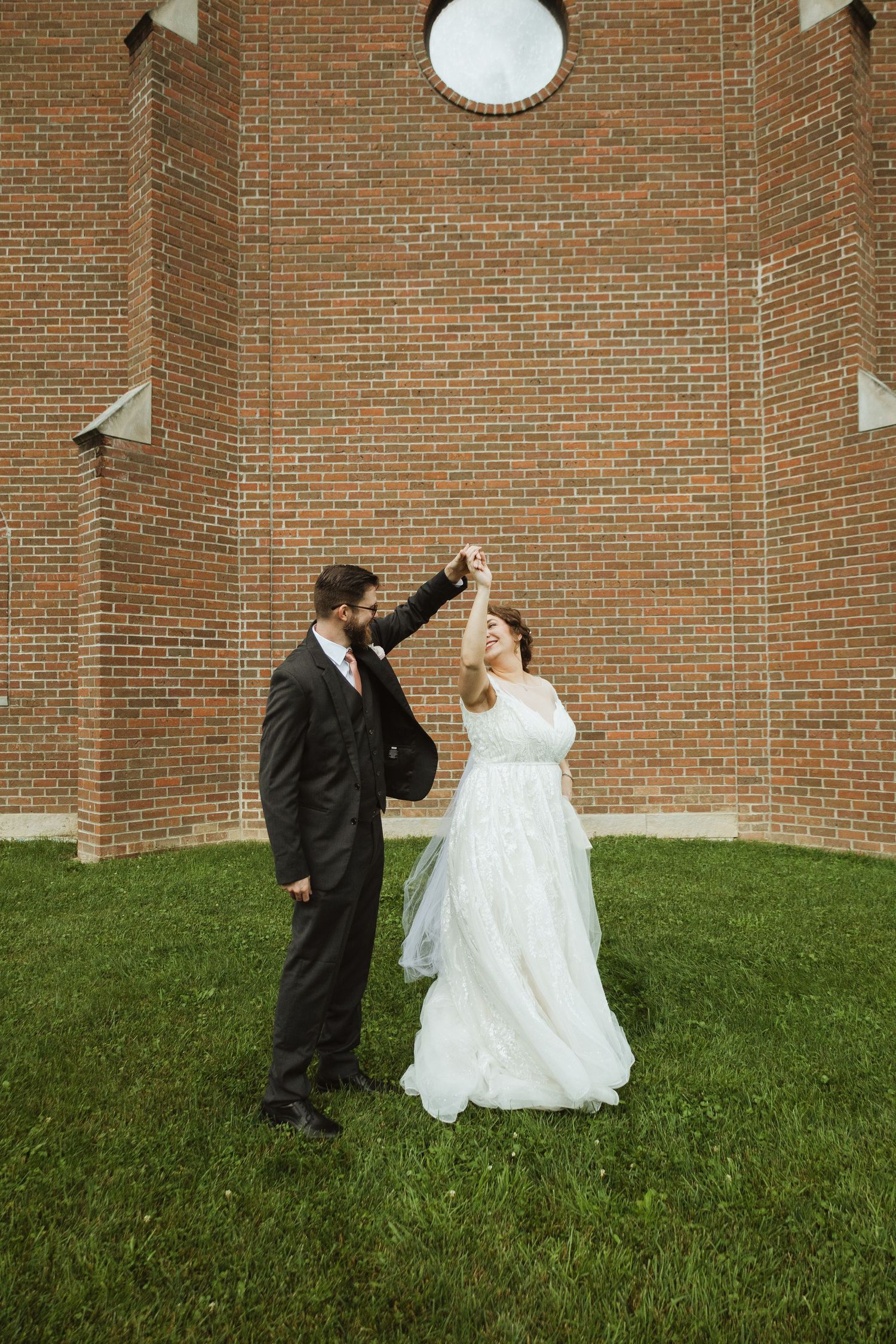 A joyful dancing moment between newlyweds outside a brick church wall.