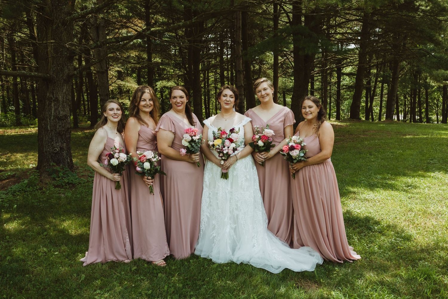 Wedding party poses in a sunlit forest setting wearing pink and white dresses.