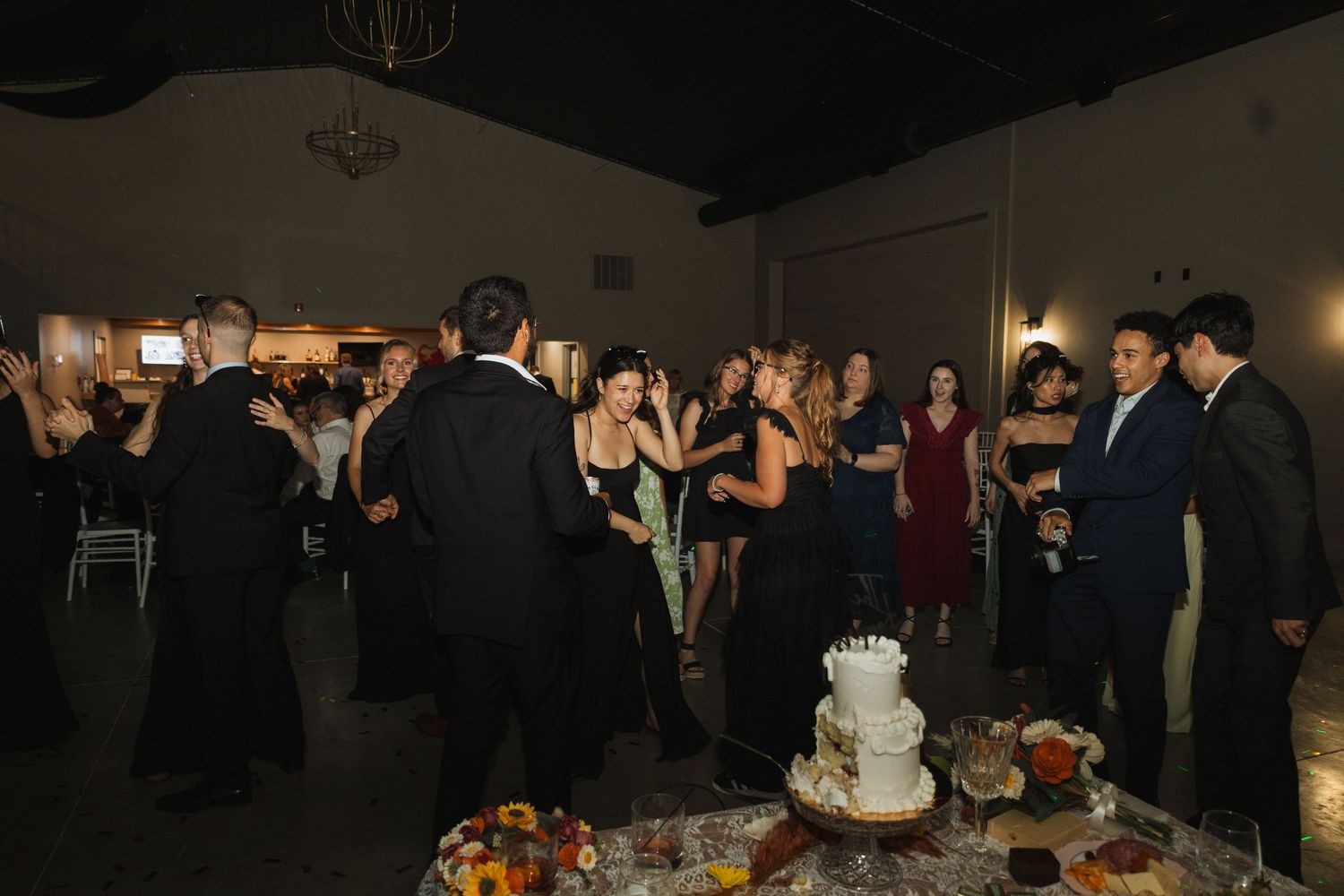 Wedding reception guests gather around a dessert table with a white tiered cake and other treats at an indoor venue.