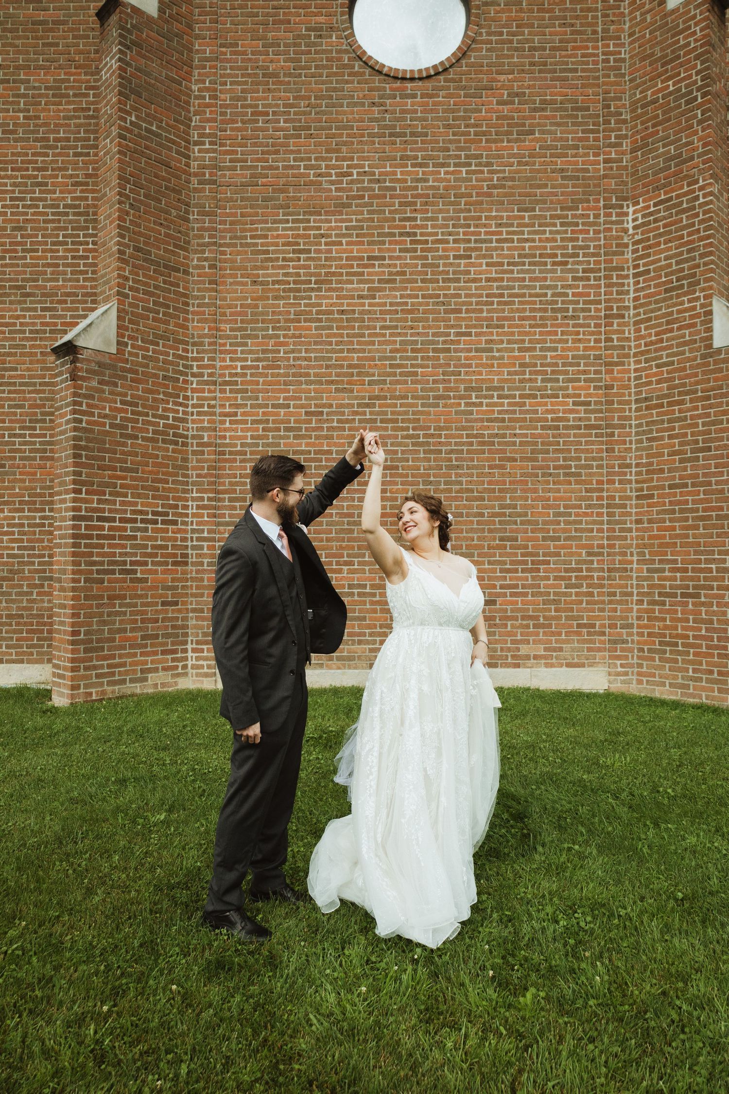 A couple dances playfully on a grassy hill against a brick wall backdrop during their wedding celebration.