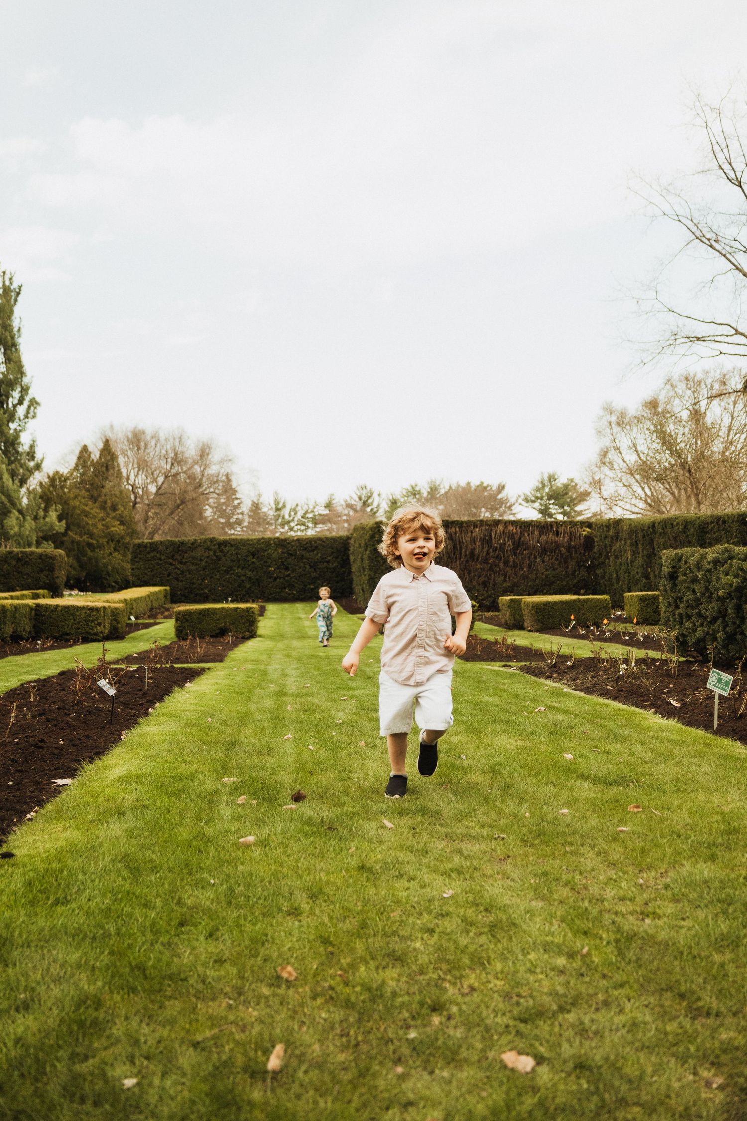 Child runs joyfully through a manicured garden with neat hedges and green lawn on a cloudy day.