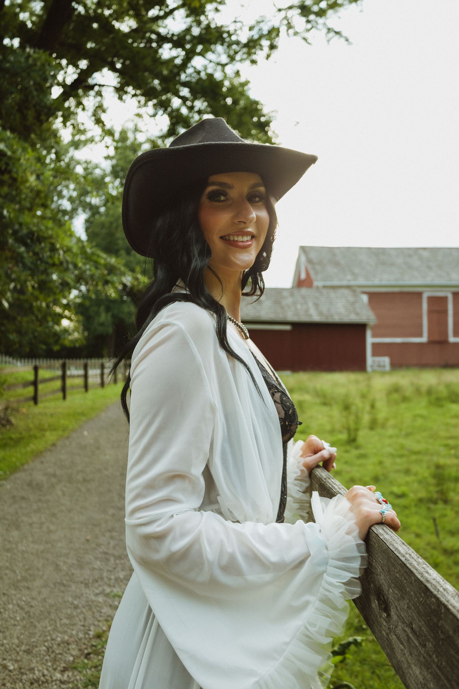 Person in elegant white attire and black hat leans against wooden fence with farmland and red barn in background.