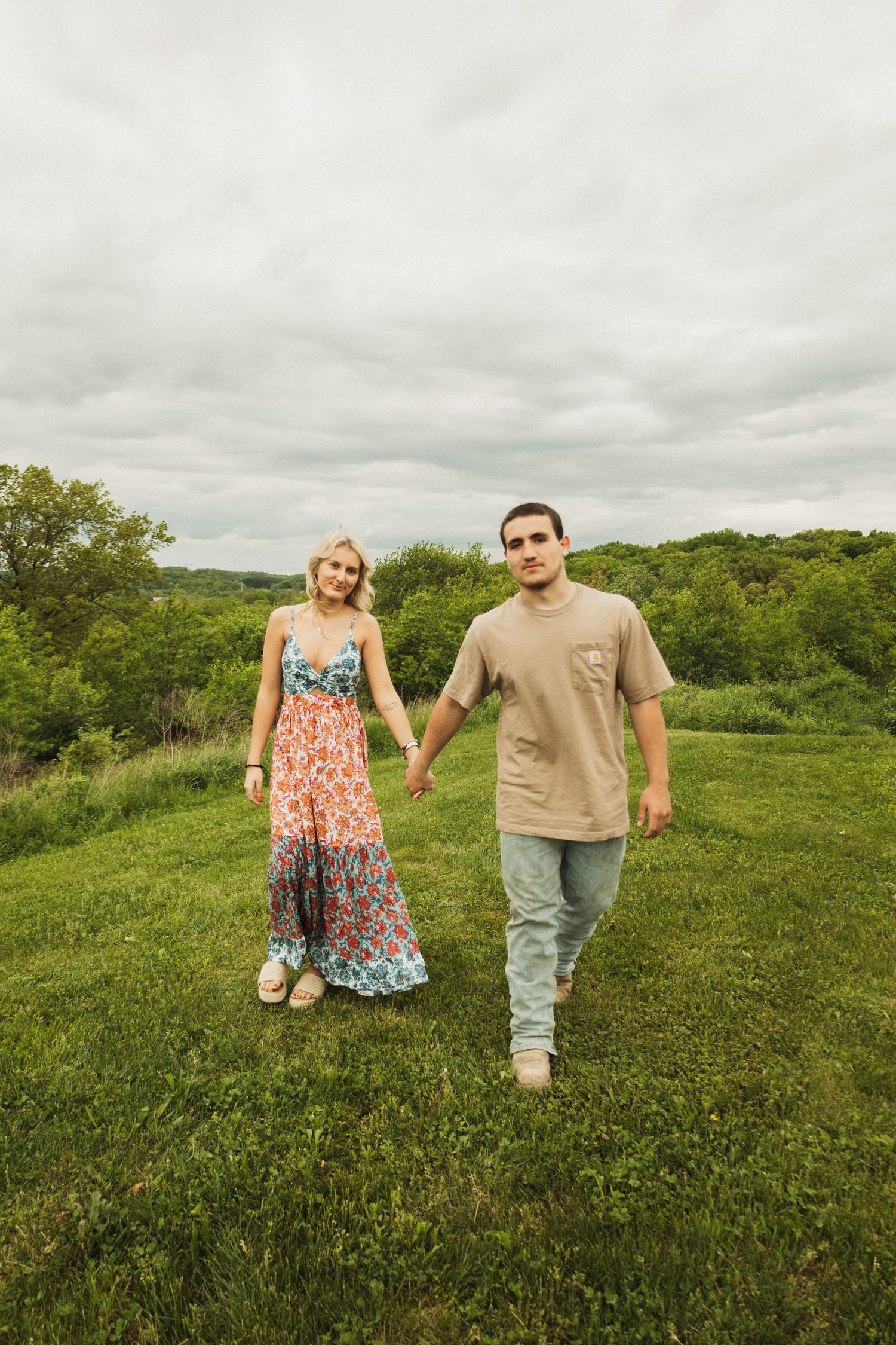 A couple walks hand in hand through a green meadow on an overcast summer day.