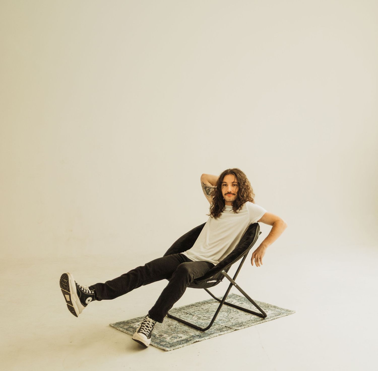 Person lounges casually in a folding chair on a patterned rug against a minimalist white backdrop.