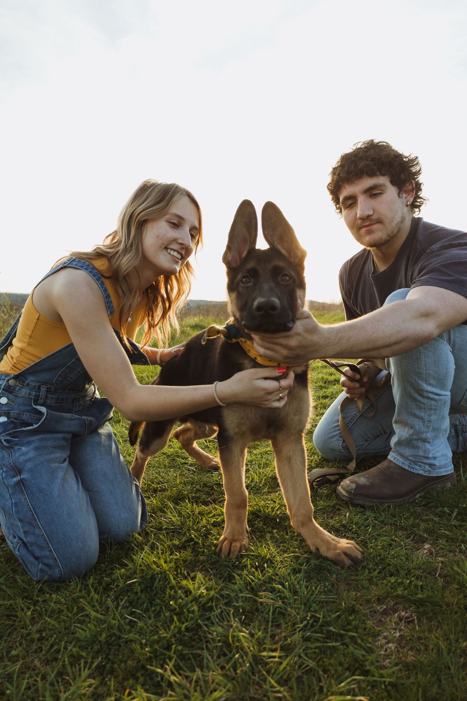 Young couple smiling and petting a German Shepherd puppy on green grass outdoors in natural light.