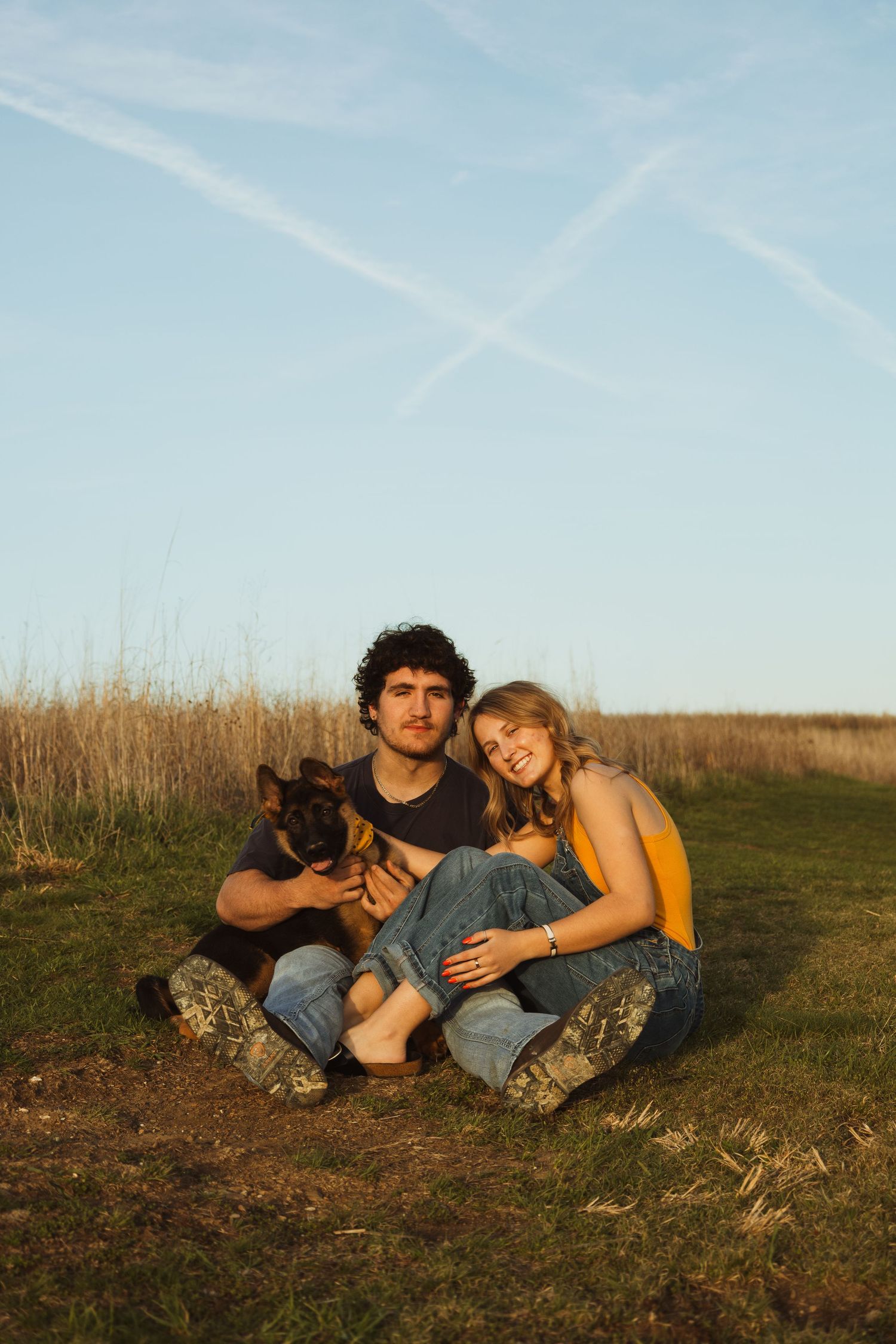 Young couple sitting on grass with German Shepherd puppy at golden hour, smiling outdoors with blue sky background.