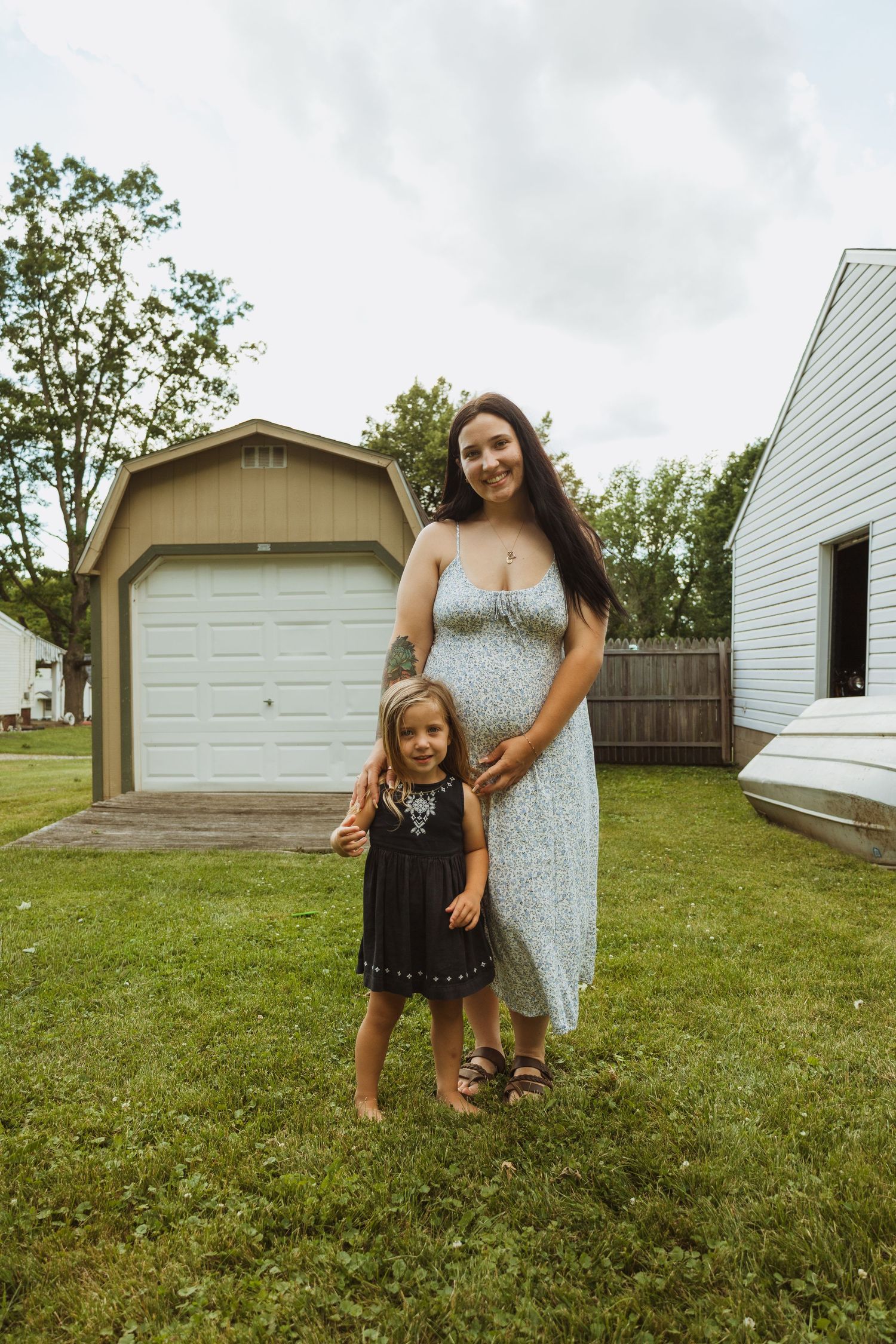 Pregnant woman in floral dress stands with young girl in black dress on green lawn near garage.