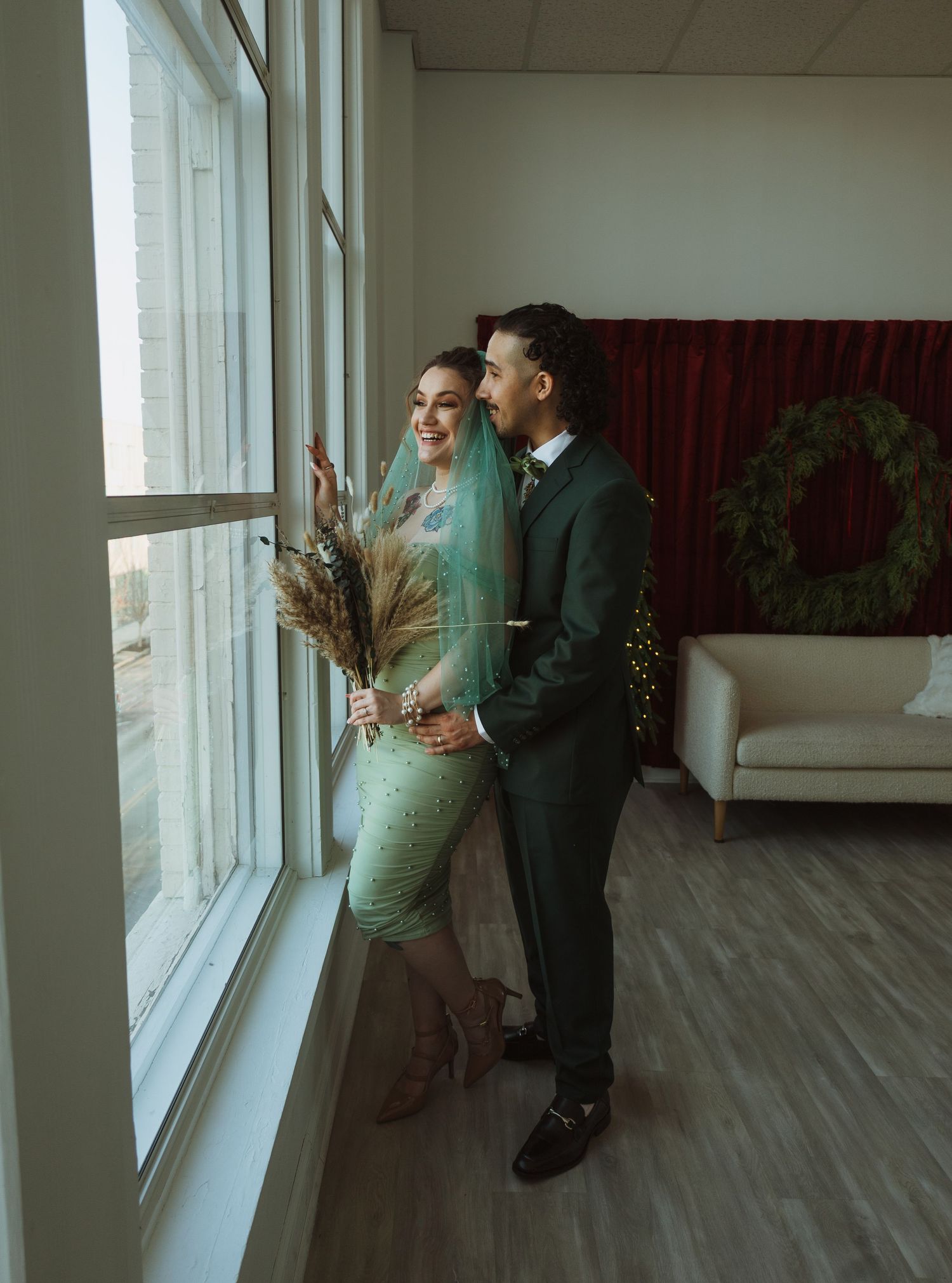 Bride in green sequin dress and teal veil holding dried flowers stands with groom by large windows.
