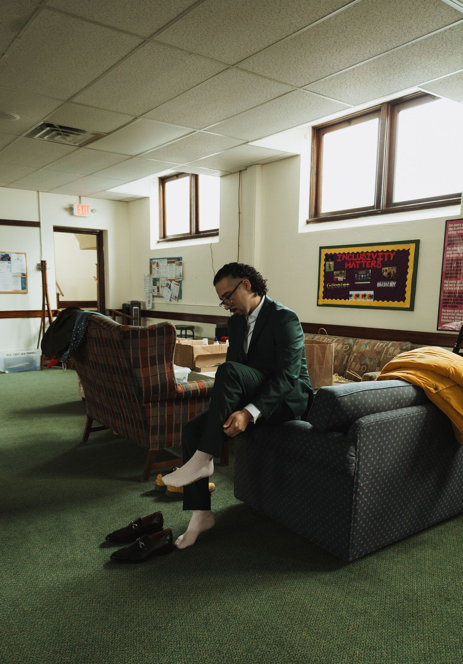 Groom in dark green suit sitting on couch adjusting his outfit in a church basement room.
