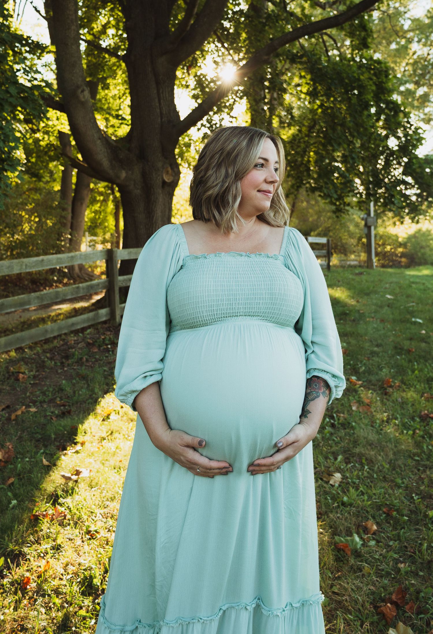 A pregnant woman in a mint green dress poses outdoors at sunset with golden sunlight filtering through trees.