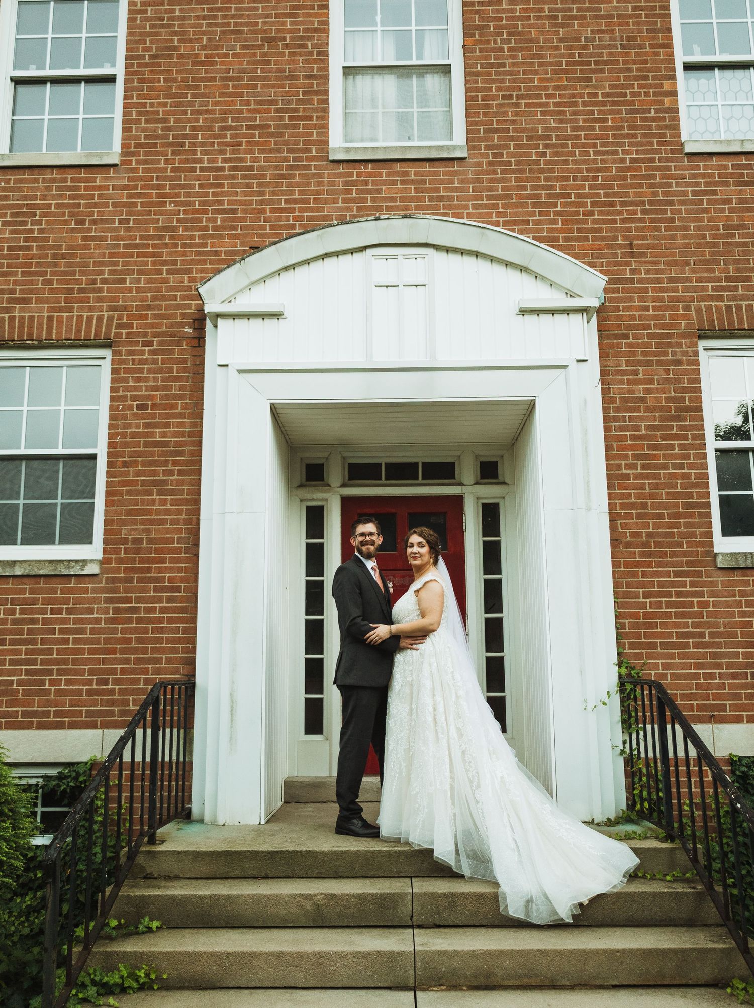 A wedding couple poses on the steps of a classic red brick building with white columns.