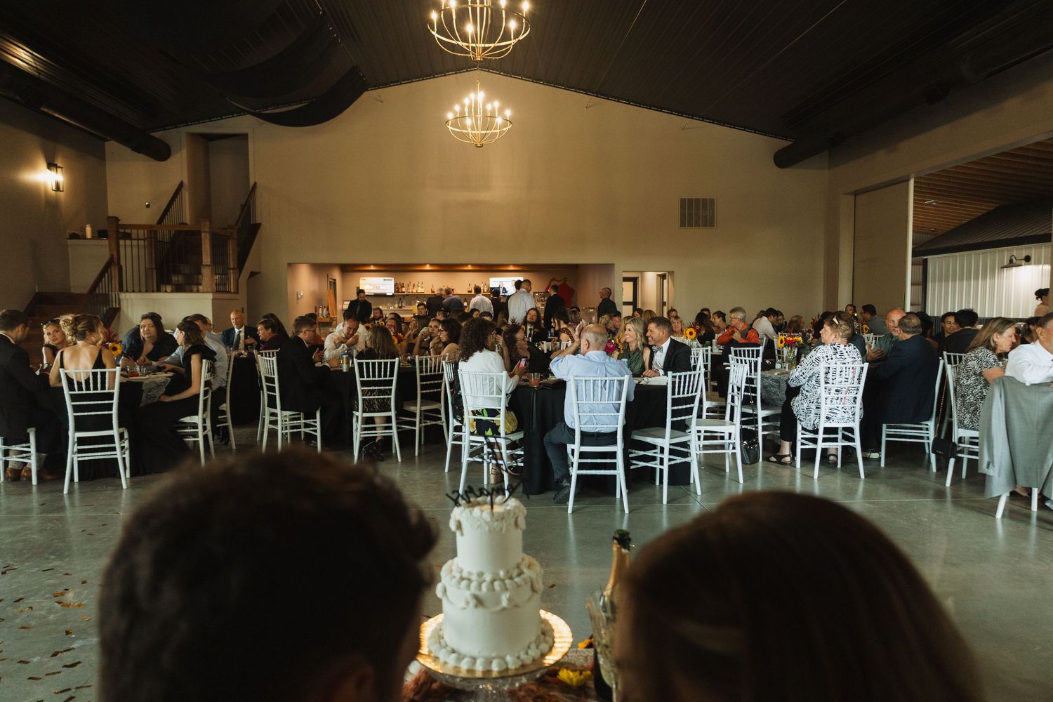 Wedding reception hall set up with white chairs and elegant chandeliers hanging from dark ceiling.
