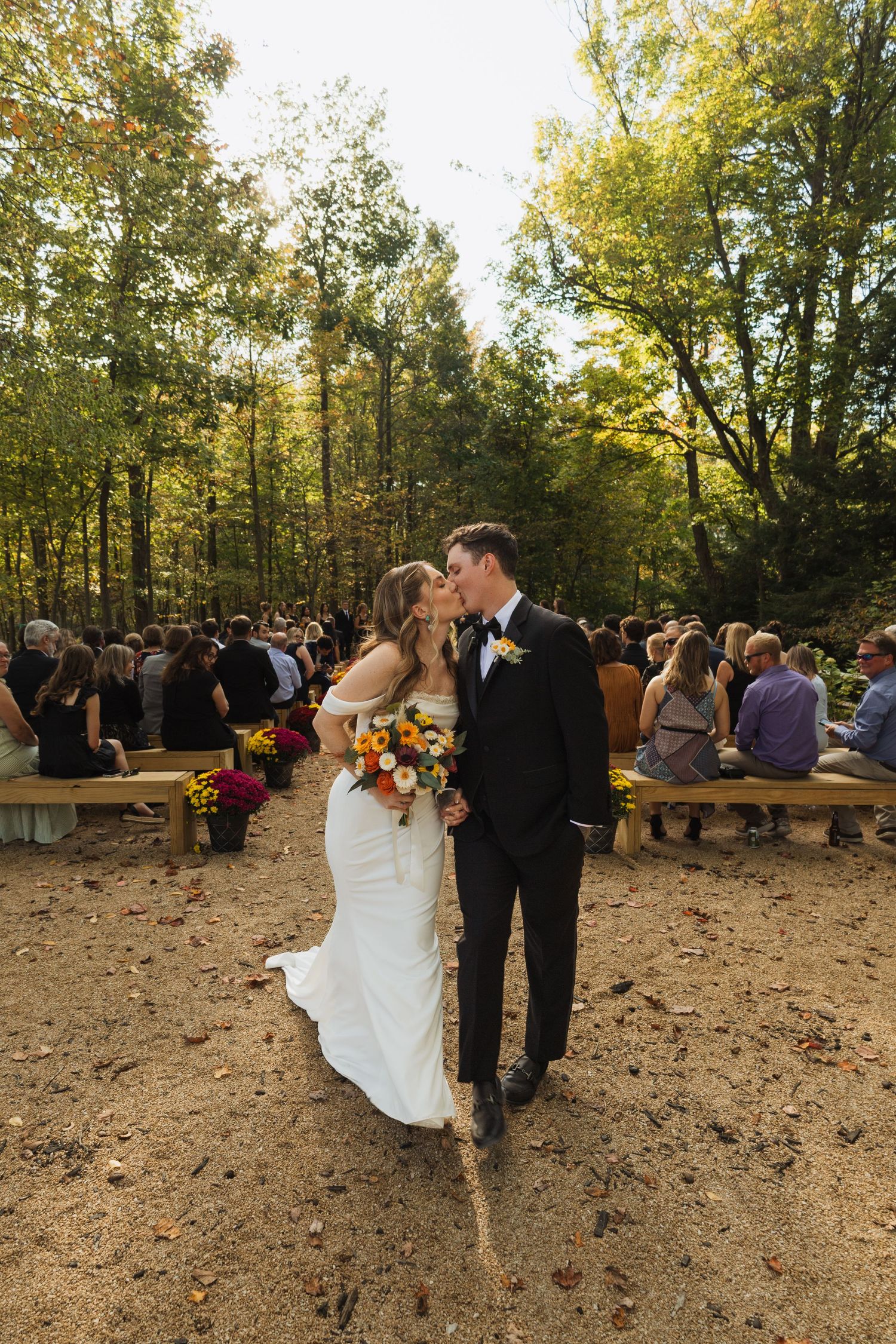 Wedding ceremony in a wooded outdoor setting with guests seated on rustic wooden benches during autumn.